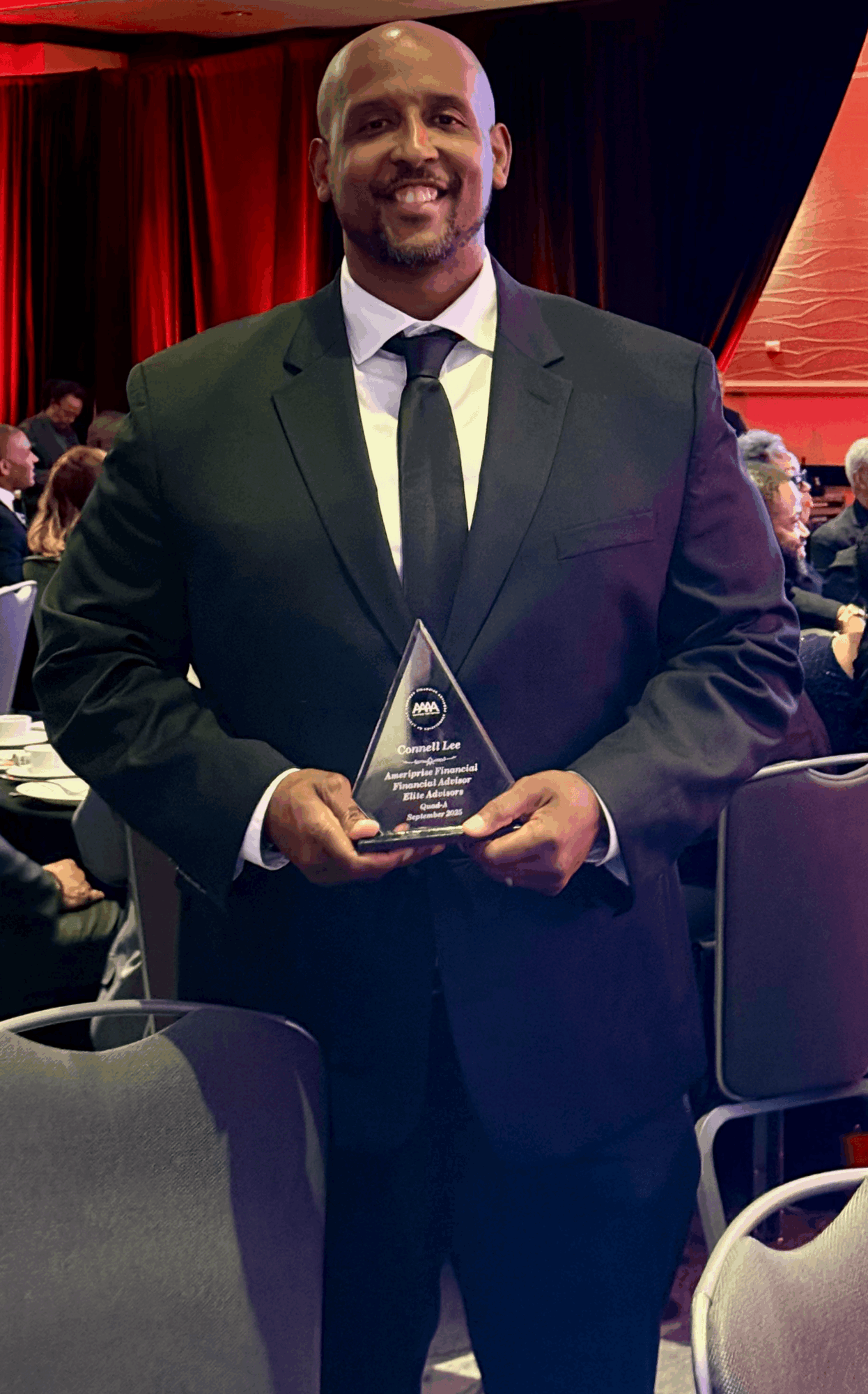 Man in a suit holding an award, smiling, at an indoor event with red curtains and seated audience.