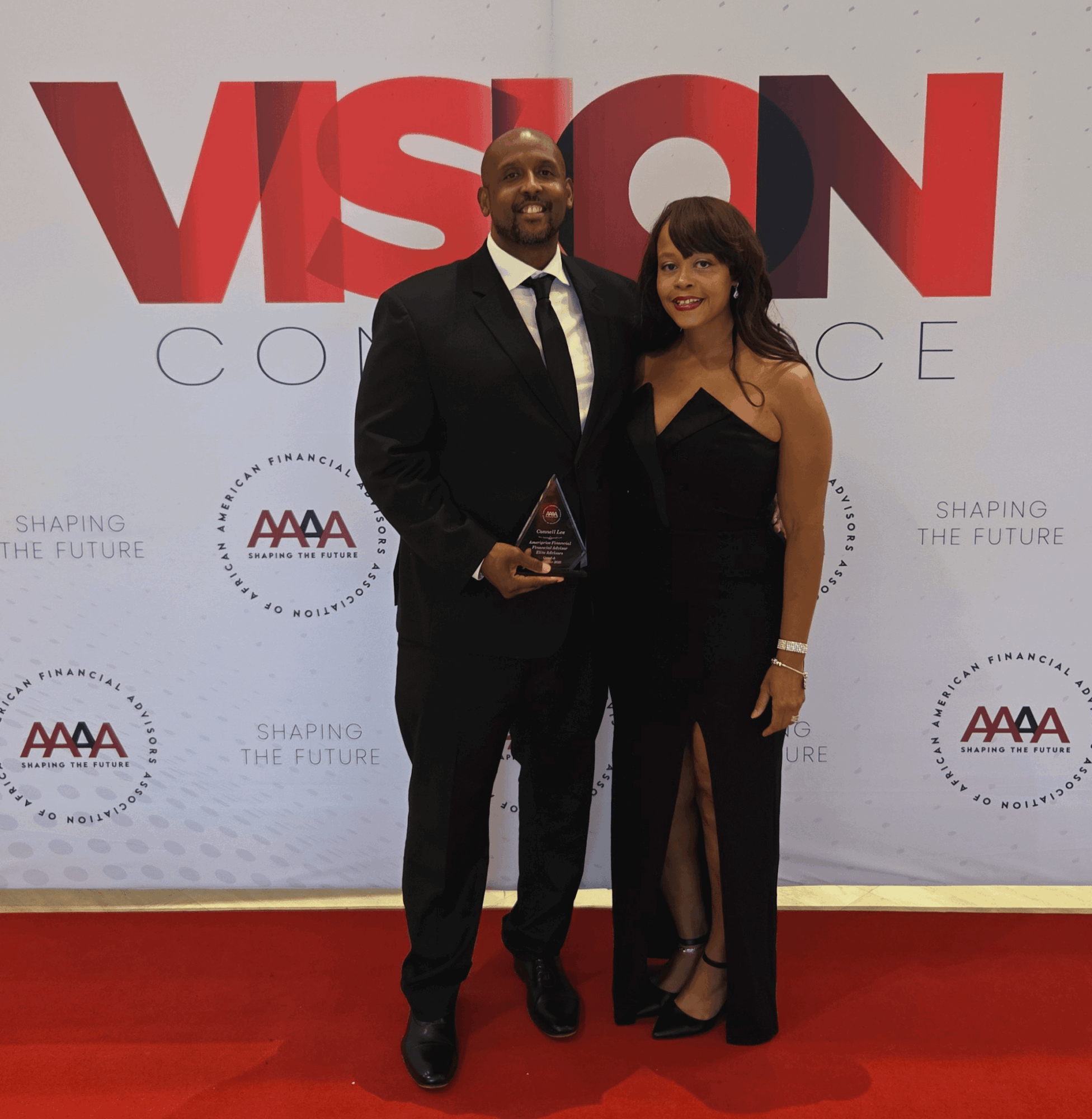 Two people in formal attire standing on a red carpet in front of a backdrop with the word 'VISION' and logos, smiling.