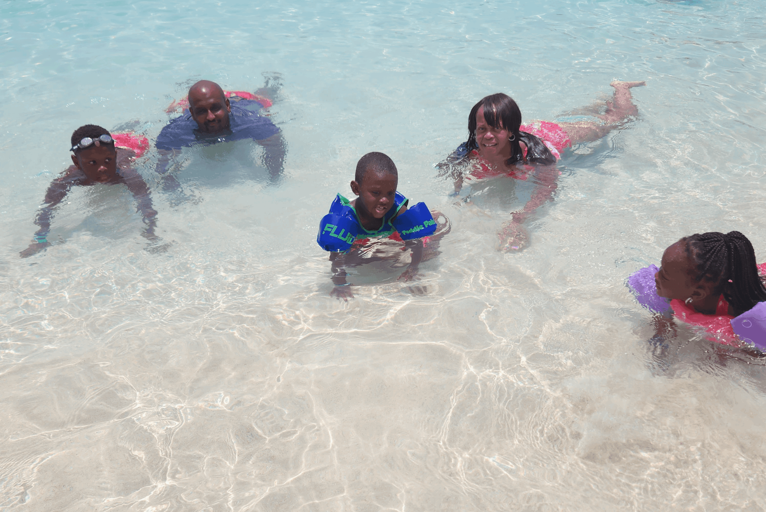 Five children and one adult in shallow water at the beach, playing and swimming, smiling and enjoying the day.