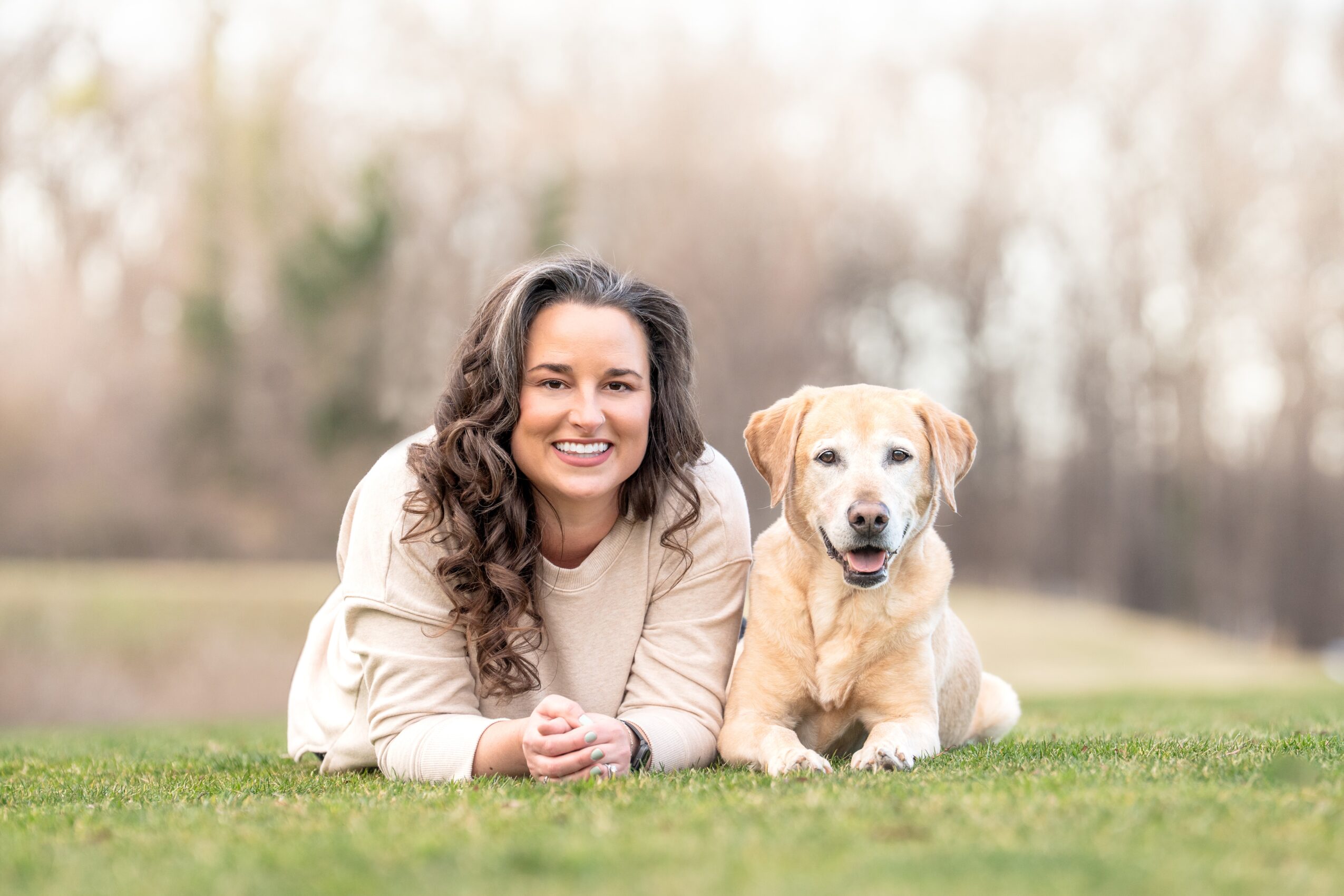 Woman lying on grass with a Labrador Retriever outdoors, trees in background, smiling, daytime