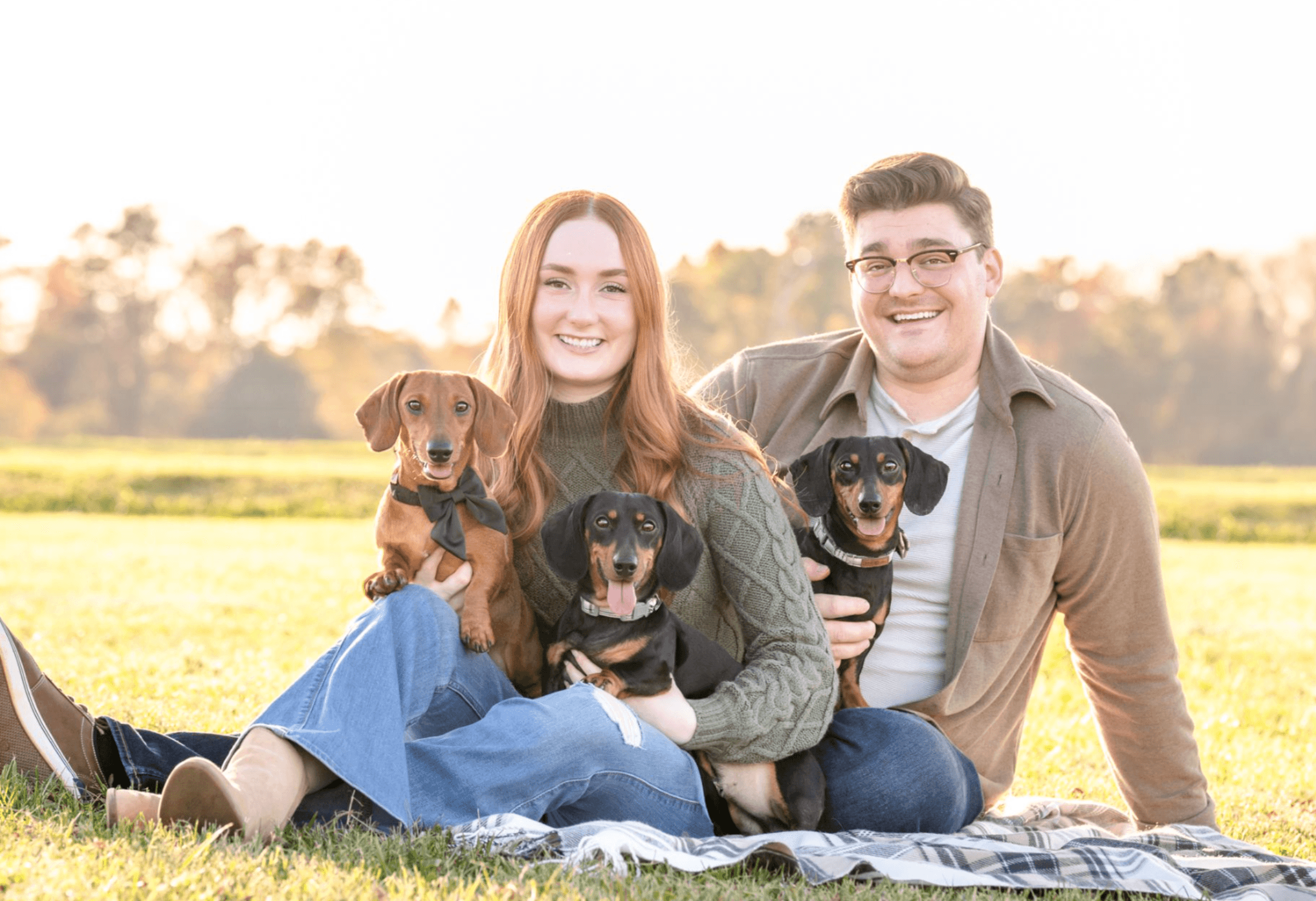 A smiling couple sitting on a blanket outdoors with three dogs, in a grassy field with trees in the background.