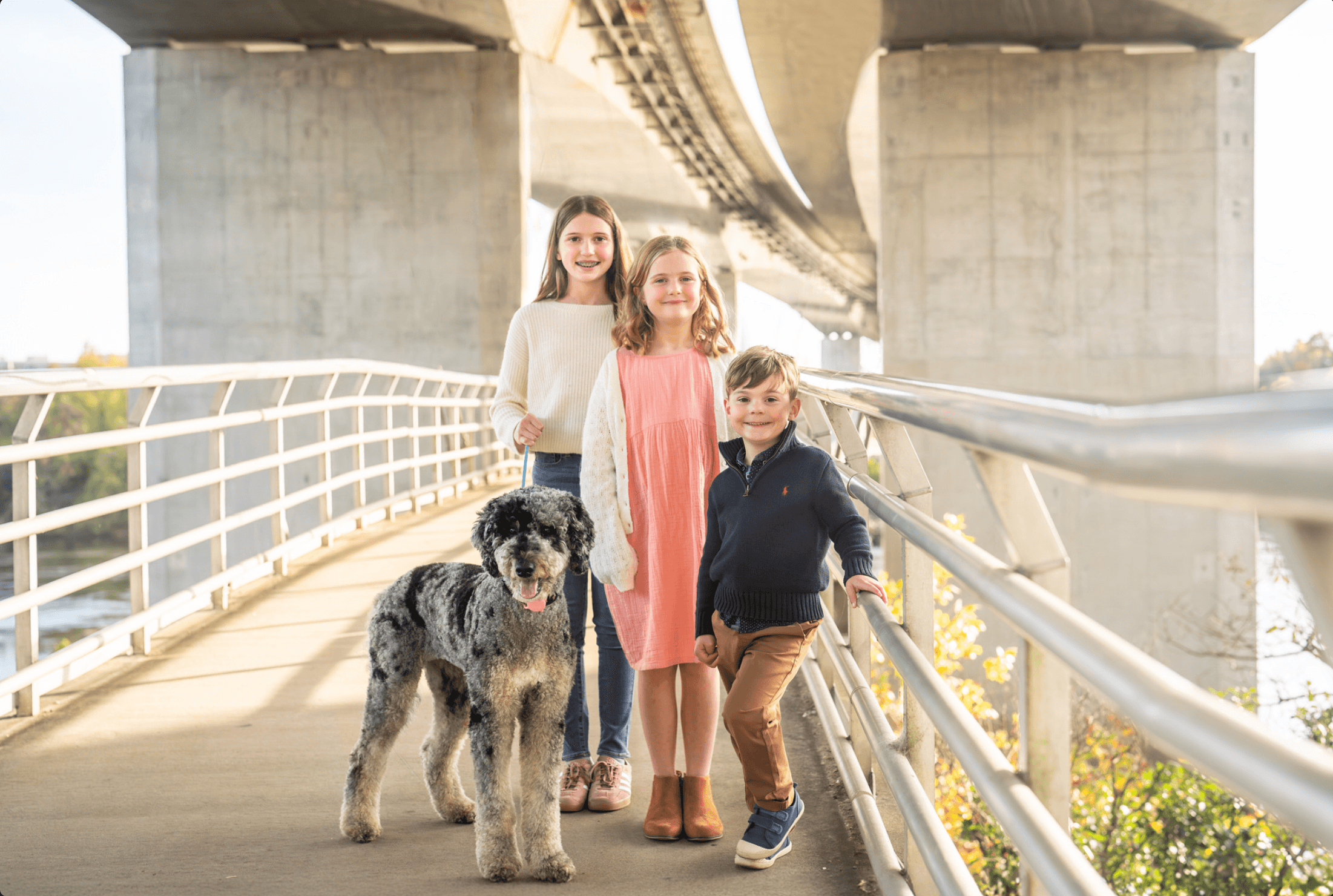 Three children and a dog standing on a bridge under a large concrete overpass, smiling at the camera.