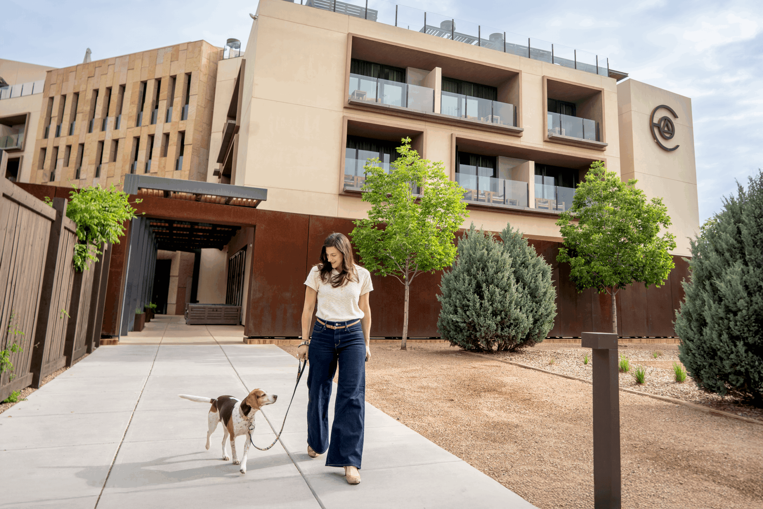 Woman walking a dog on a leash outside modern building with trees and bushes nearby.