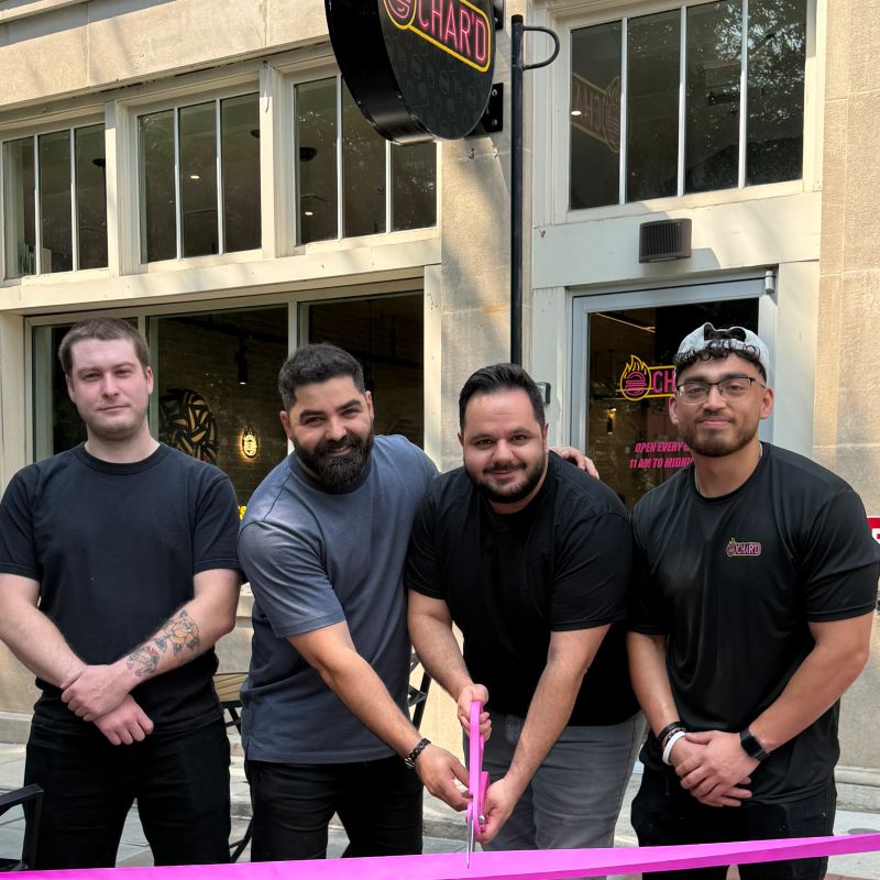 Four men standing outside a building with large windows, one holding a pink ribbon, celebrating an event.