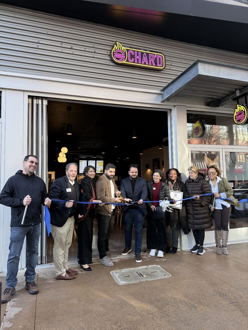 Group of people standing outside a store, holding a ribbon for a ribbon-cutting event, with a store sign above.