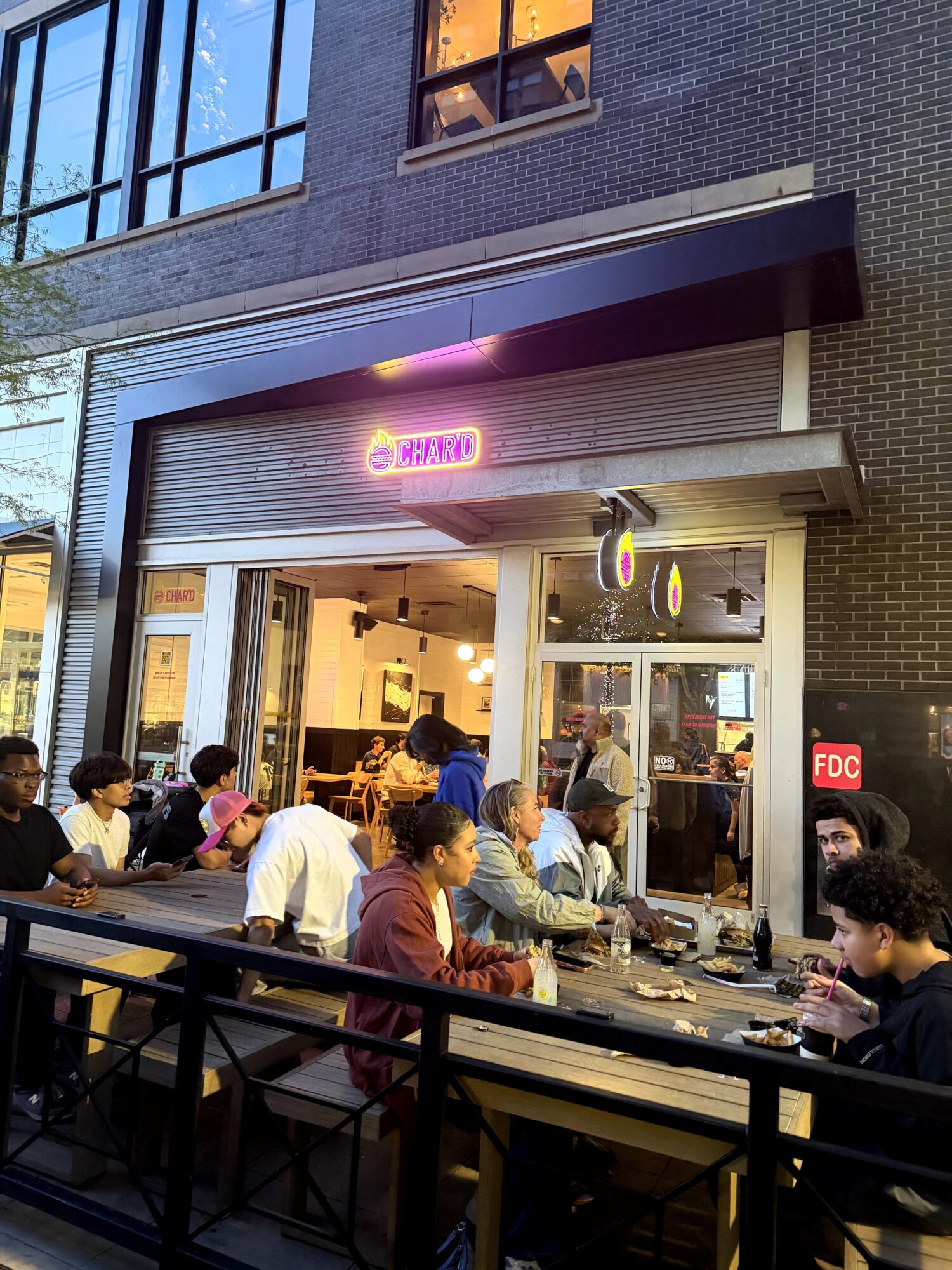 People sit at tables outside a restaurant with a neon sign, large windows, and a brick facade, during evening.