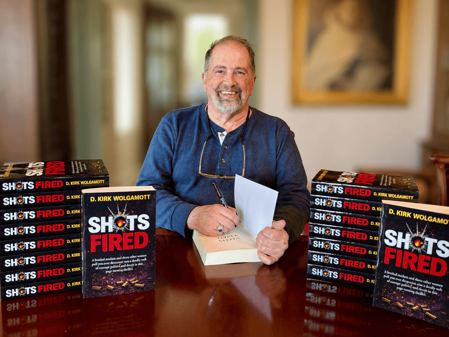 A smiling man with glasses and a beard sits at a table with stacks of books titled 'Shots Fired'.