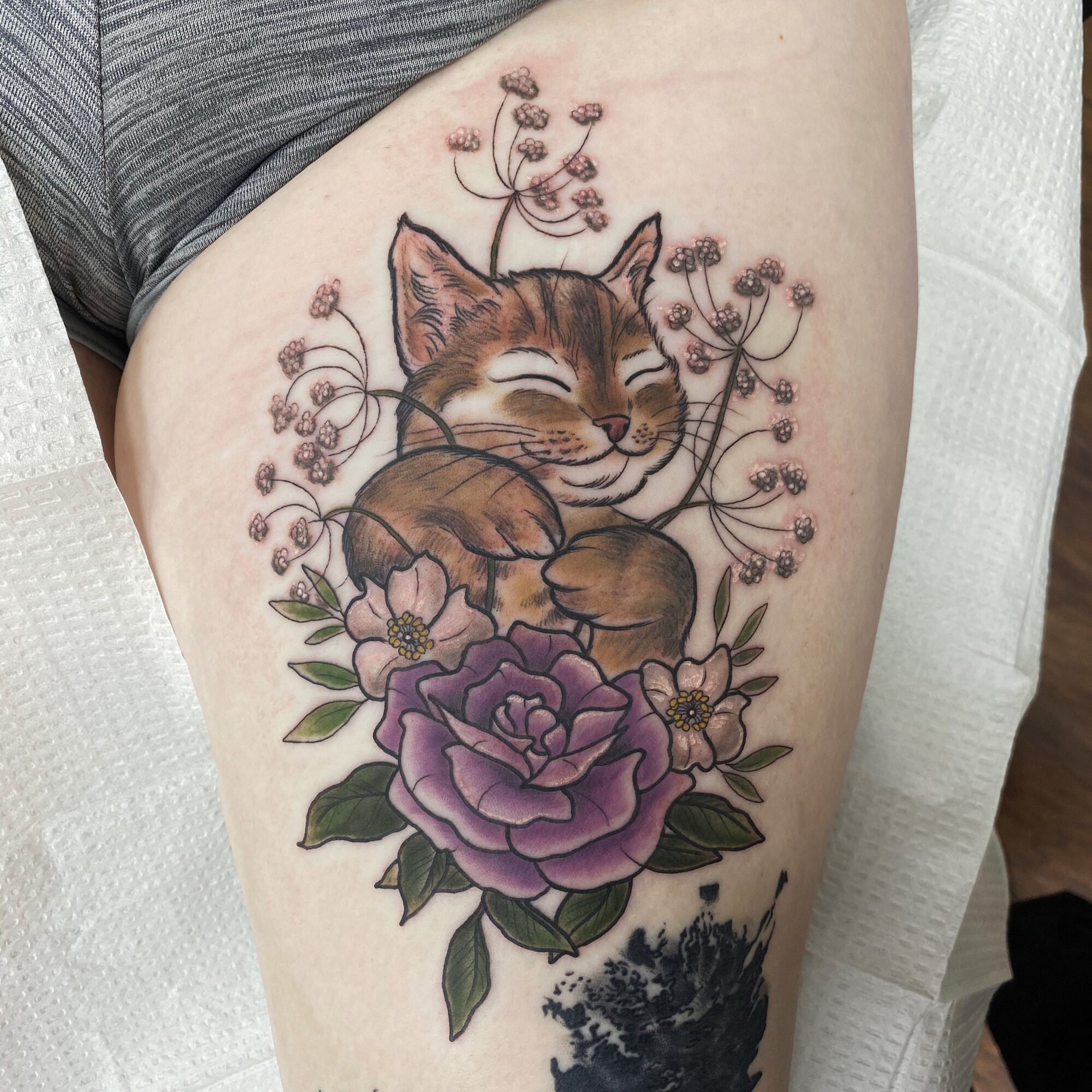 Cat with closed eyes surrounded by purple and pink flowers, detailed and colorful tattoo on thigh.