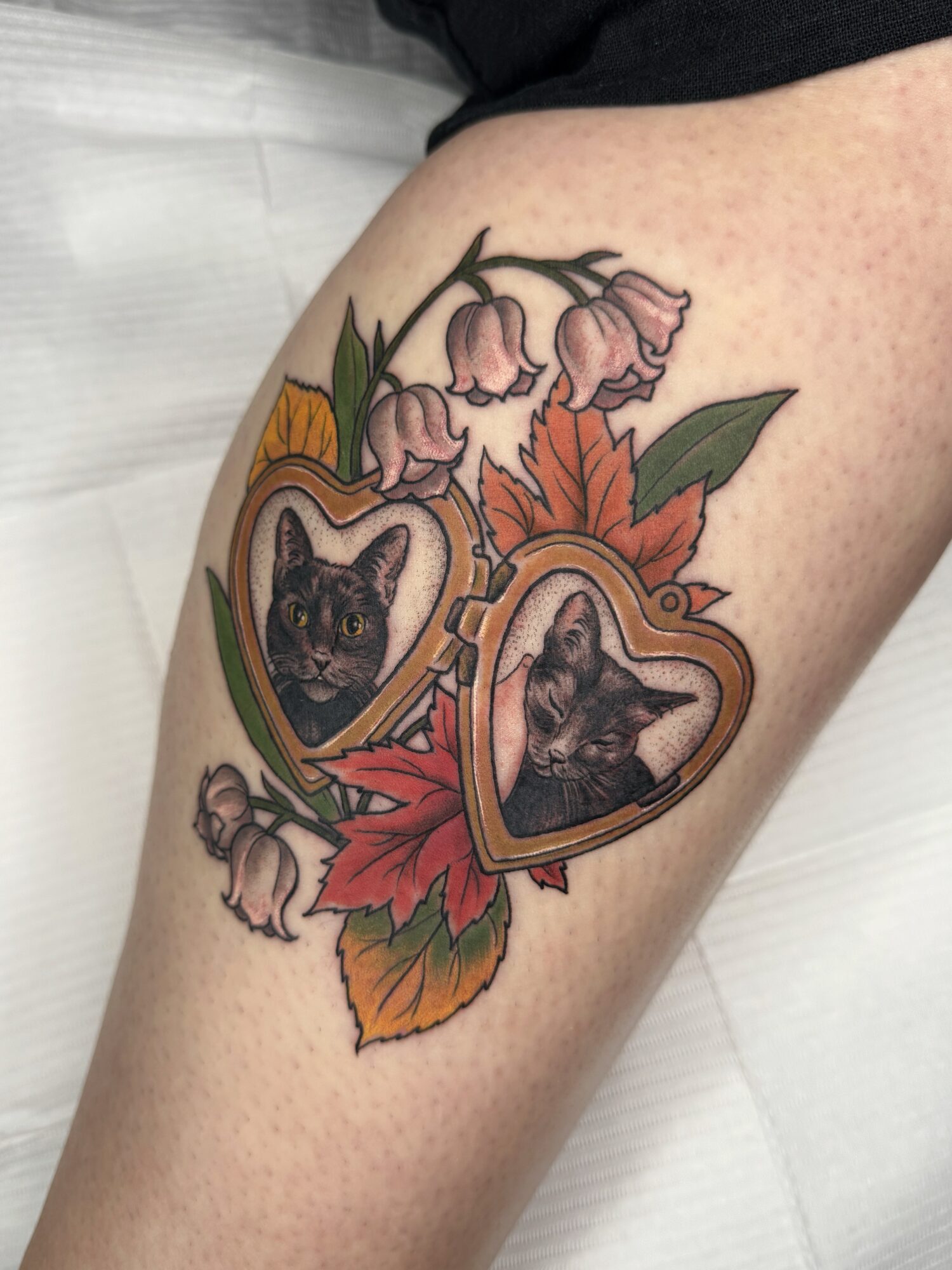 Tattoo of two black cats in heart-shaped frames surrounded by pink flowers, orange leaves, and green foliage on skin.