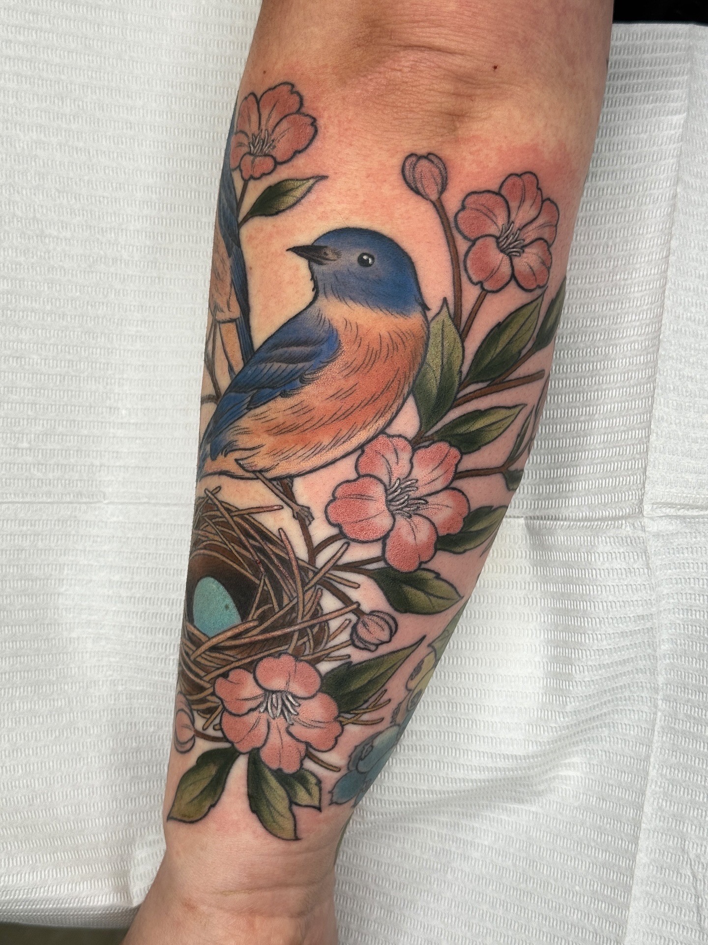 Colorful bird tattoo surrounded by pink flowers and green leaves on person's arm.