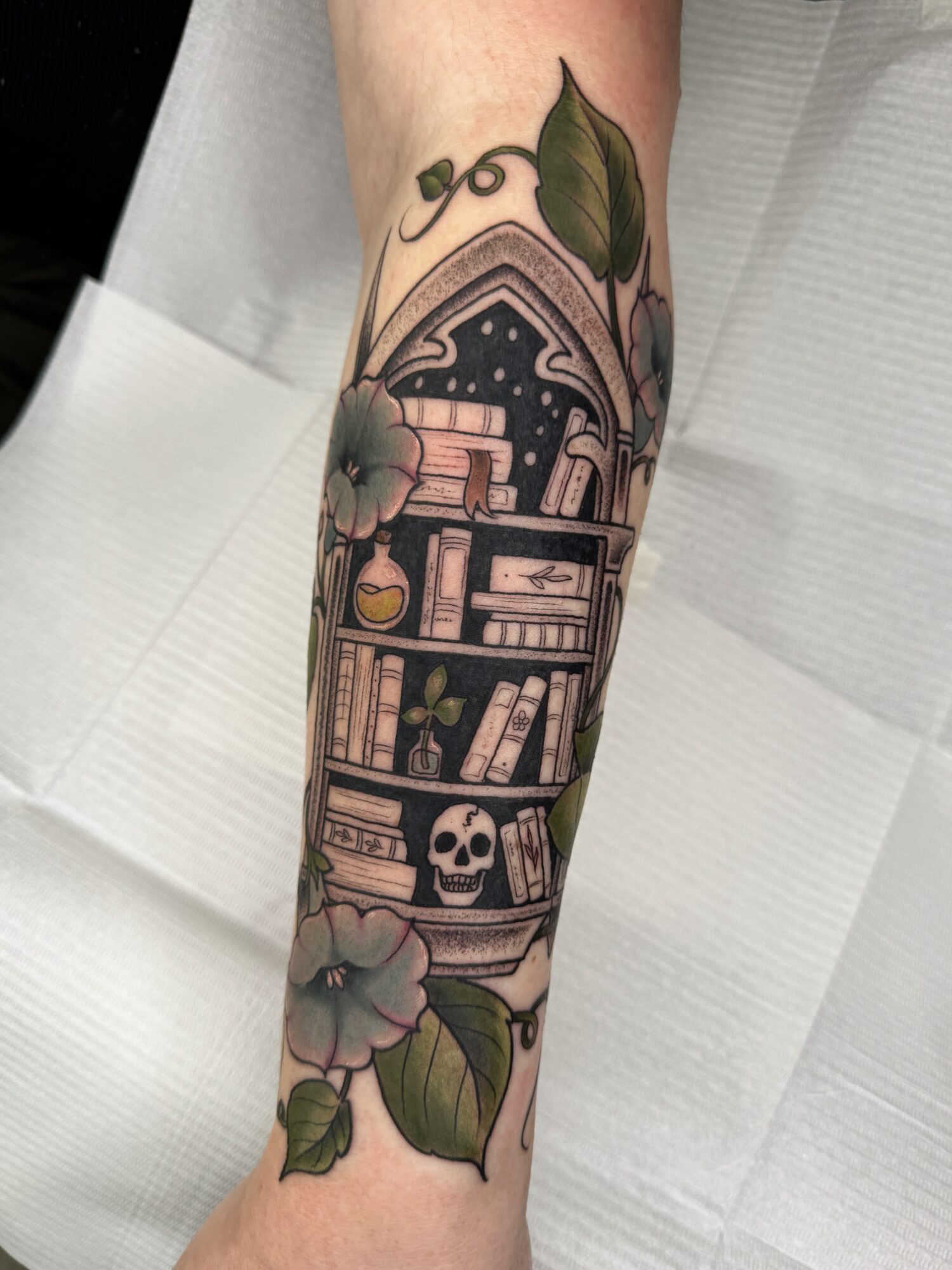 Tattoo of a bookshelf with a skull, flowers, and leaves on a person's arm.