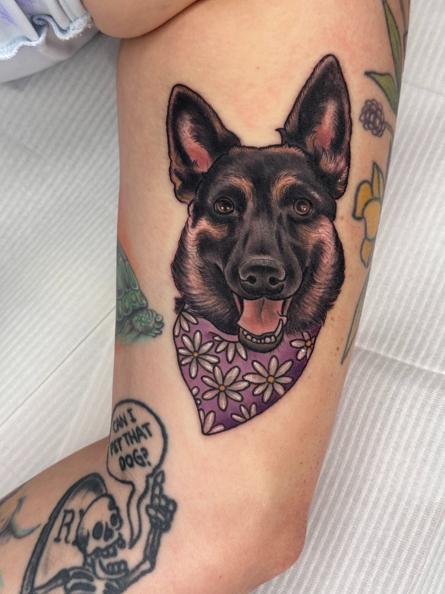 Tattoo of a dog's face with a purple floral band around its neck on a person's arm.