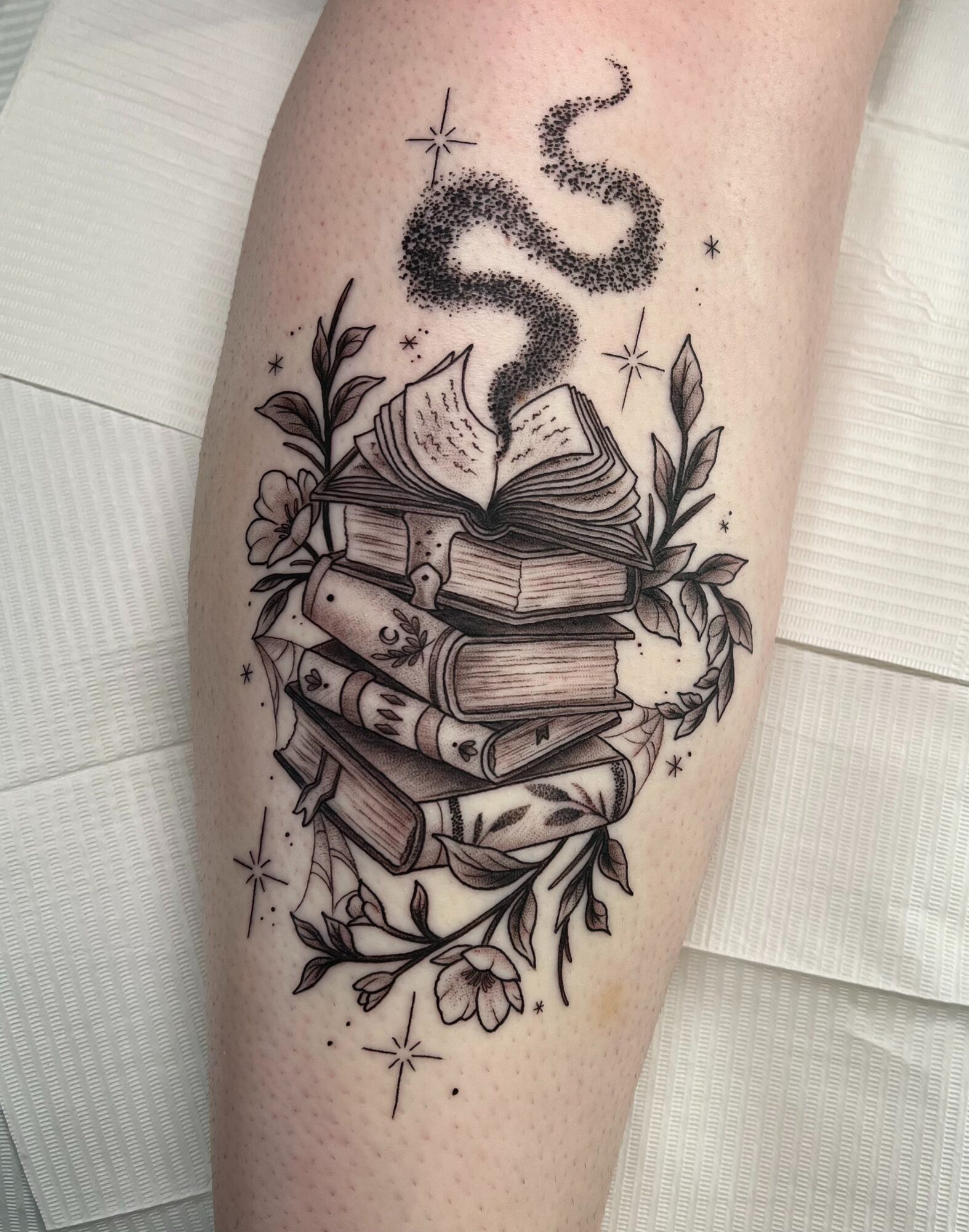 Tattoo of a stack of books with a snake emerging from the top, surrounded by leaves and stars, in black ink.