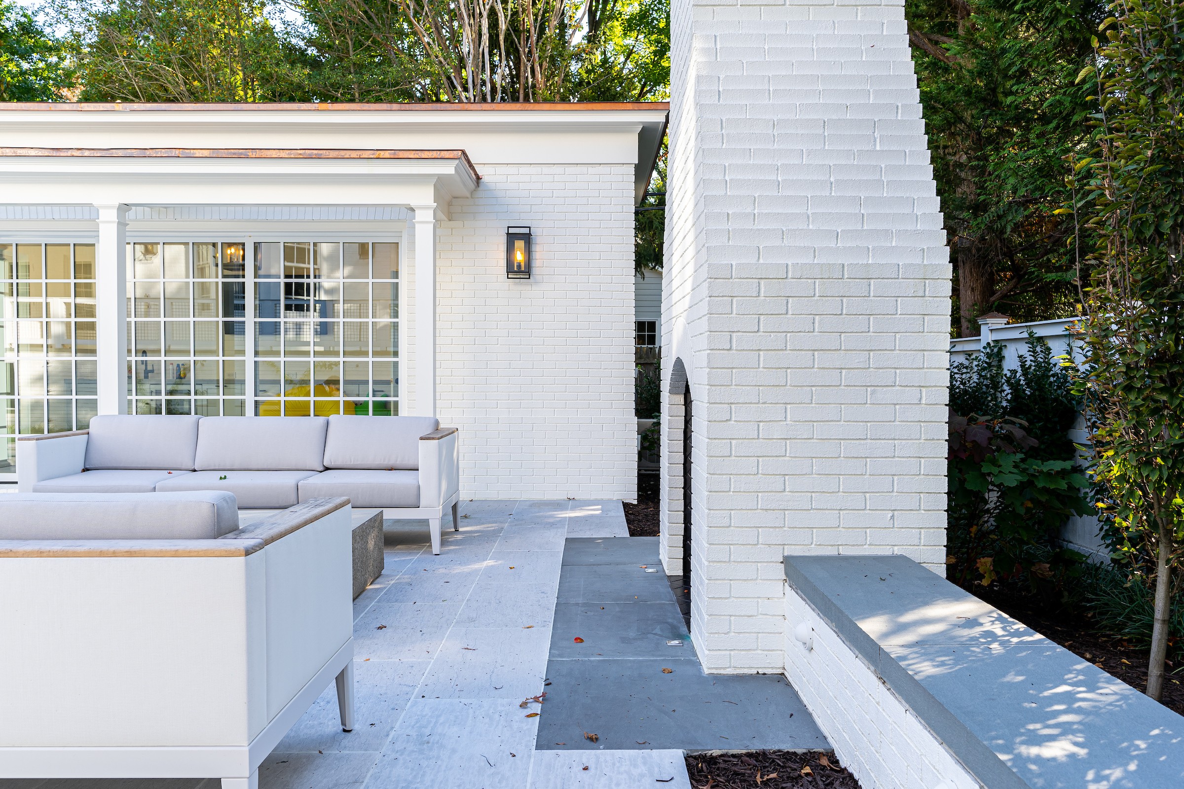 Outdoor patio with white furniture, brick fireplace, and a large window, surrounded by greenery.