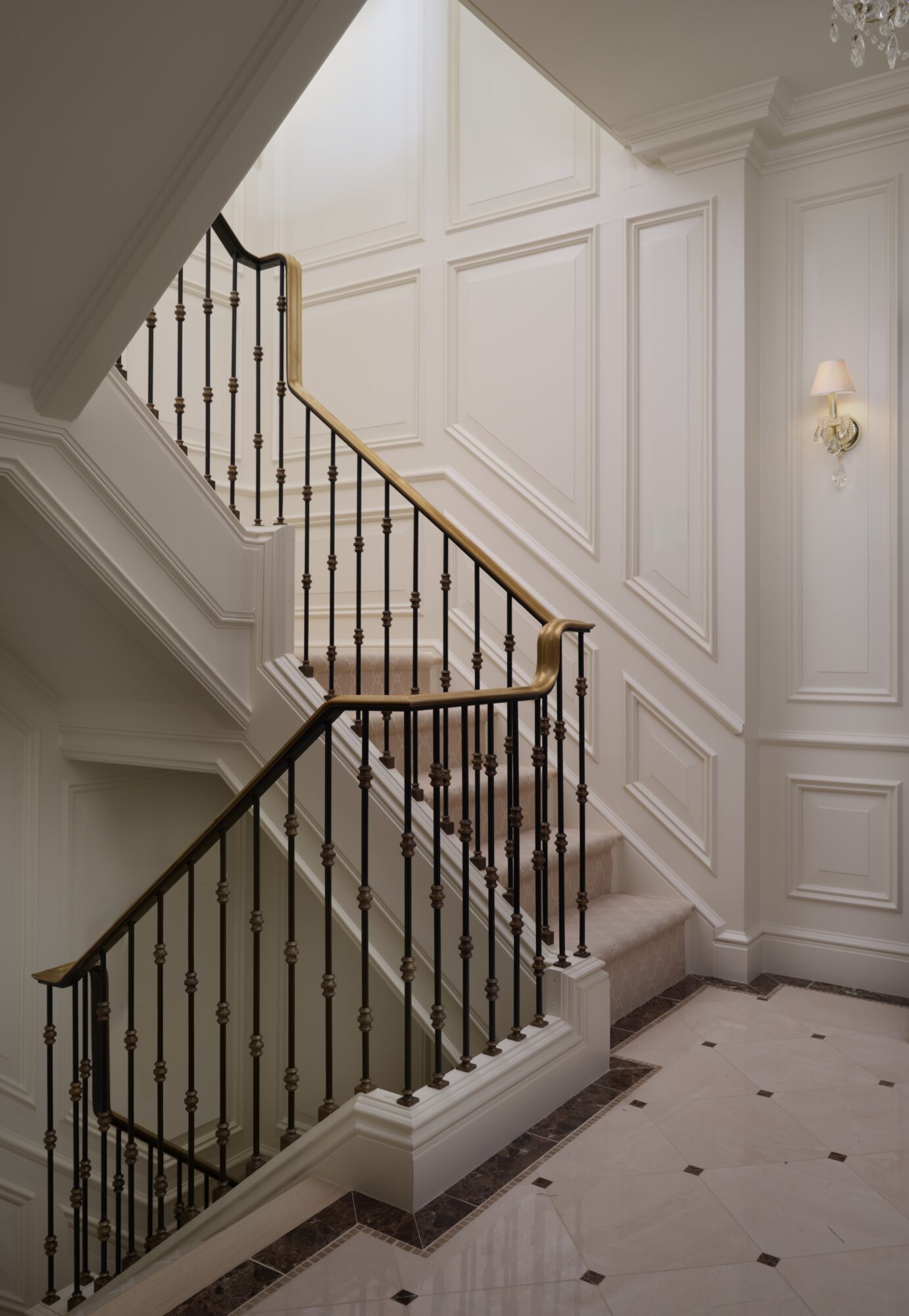 Interior staircase with white paneled walls and black and gold railing, ascending to upper floor, elegant lighting fixtures visible.