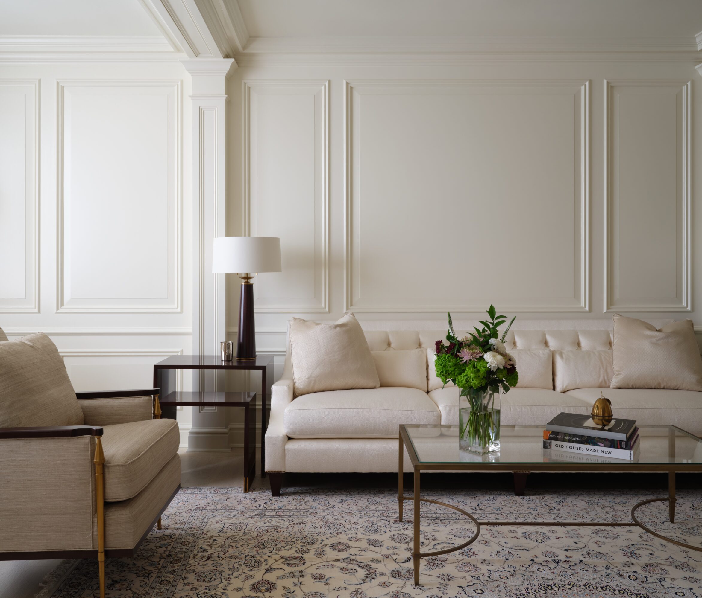 Living room with white paneled walls, beige sofa, side table with lamp, and a coffee table with flowers and books.