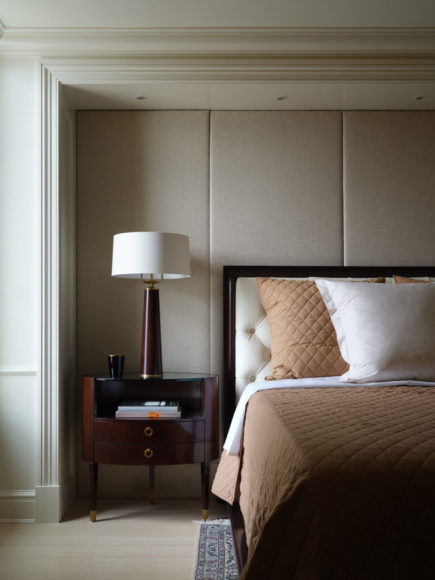 Part of a bedroom with a bed, beige headboard, brown pillow, white pillow, brown bedspread, and a nightstand with a lamp and books.