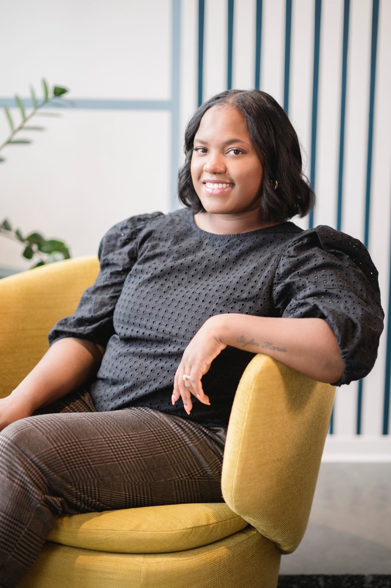 Woman with shoulder-length hair sitting on a yellow chair, smiling, in a modern room with vertical blue and white stripes.