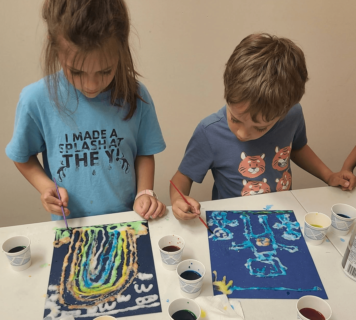 Two children painting abstract pictures at a table with cups of paint, focused on their artwork.
