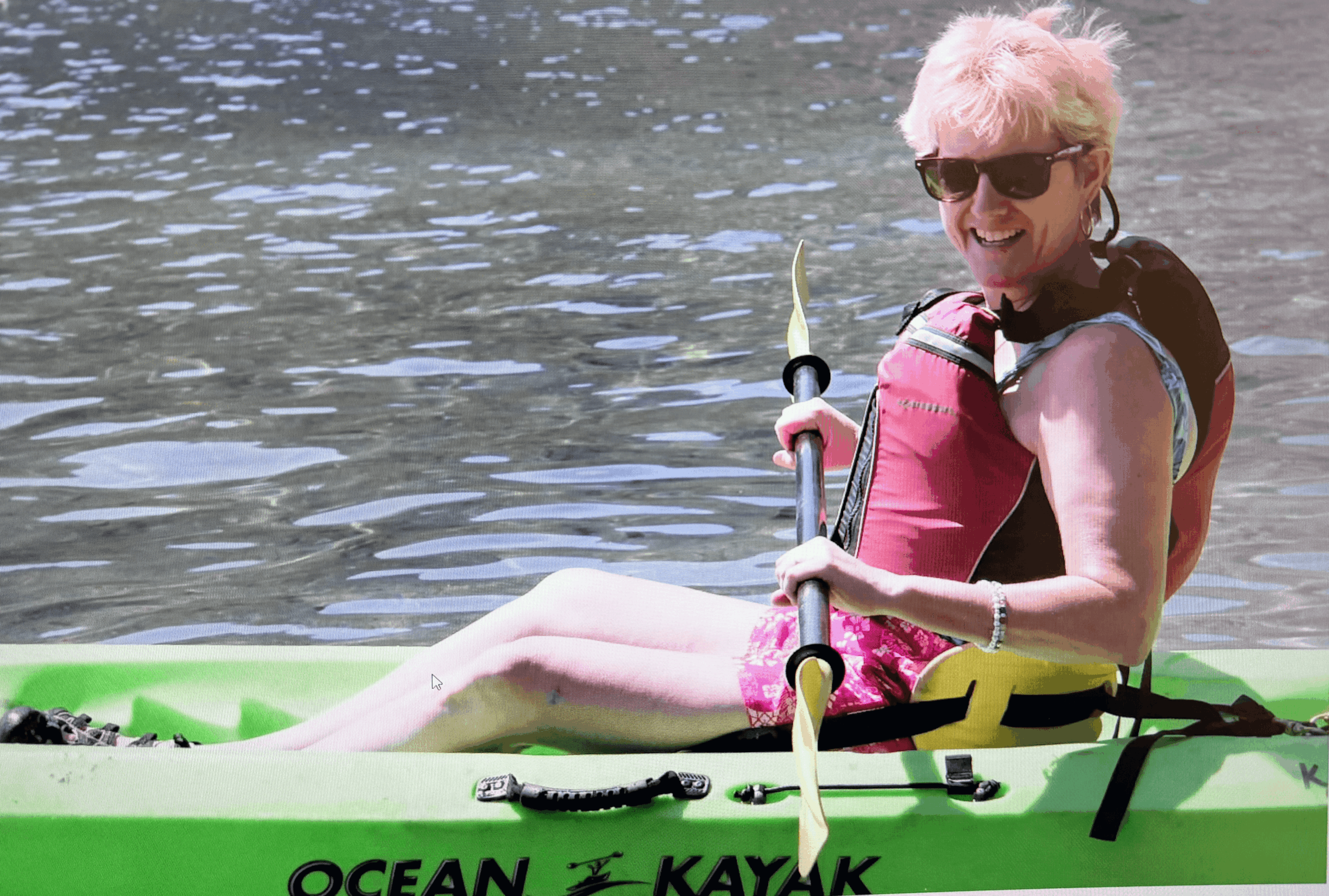 Woman with short blonde hair wearing sunglasses and a life jacket, sitting in a kayak on water, smiling.