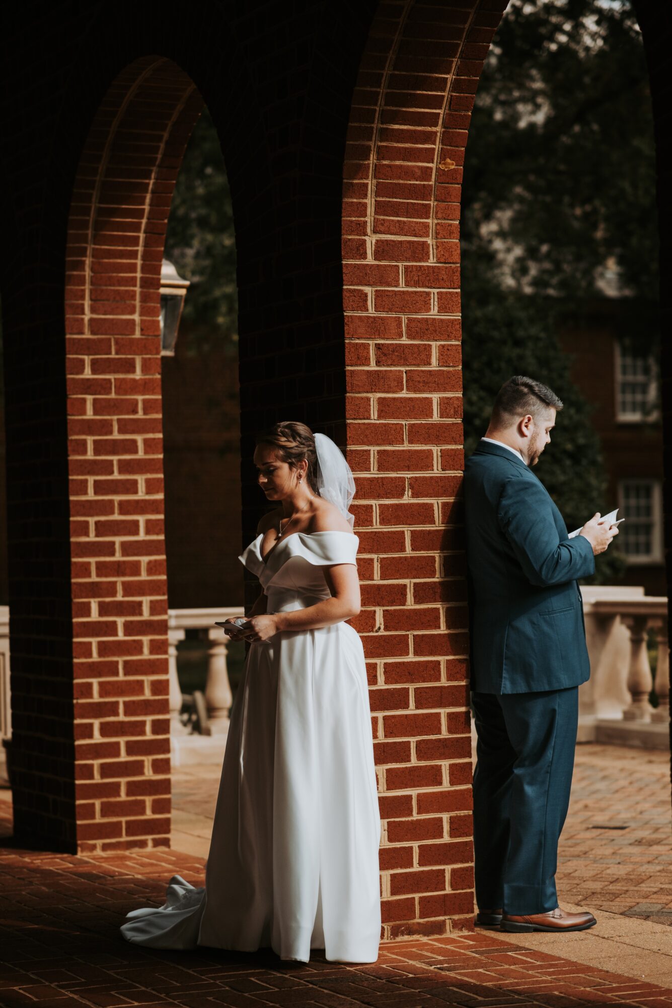 Bride in a white dress and veil, and groom in a suit, stand apart under brick arches outdoors.