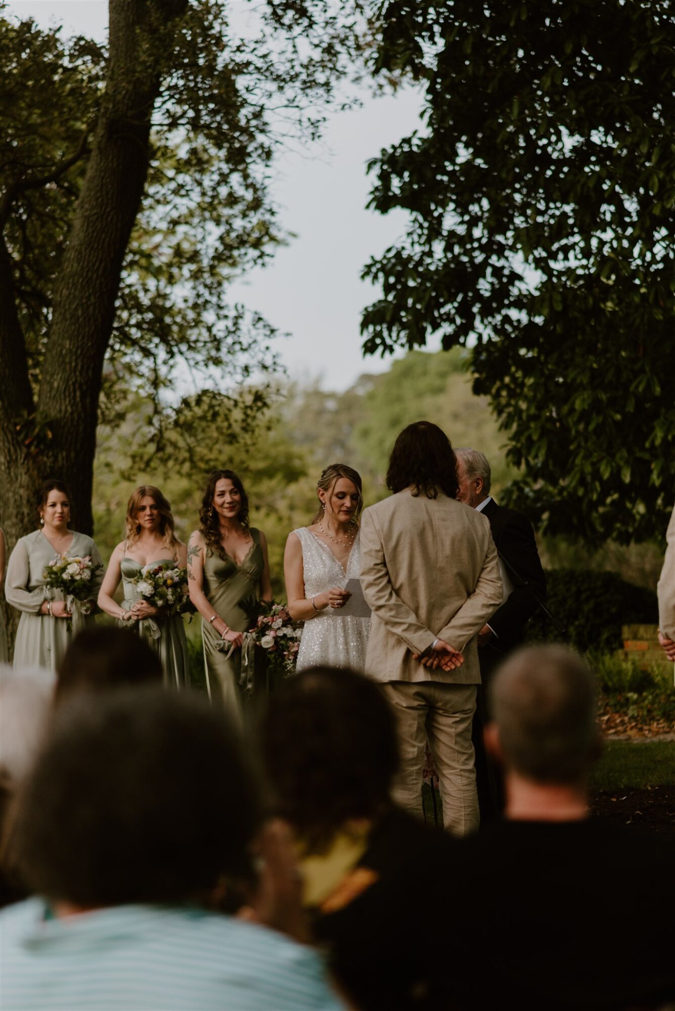 People gathered outdoors under trees, with a person in a white dress and others in formal attire, facing a person in a beige suit.