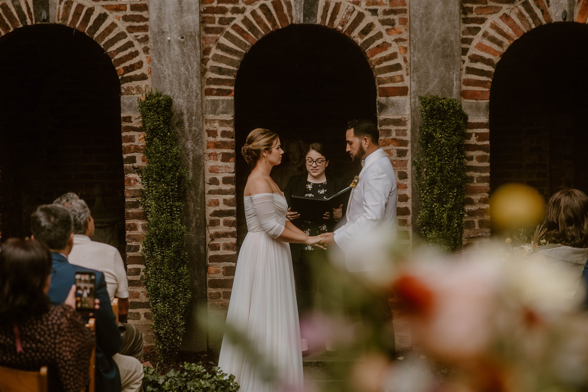 Bride and groom exchange vows in front of officiant during wedding ceremony, guests seated, brick arches in background.