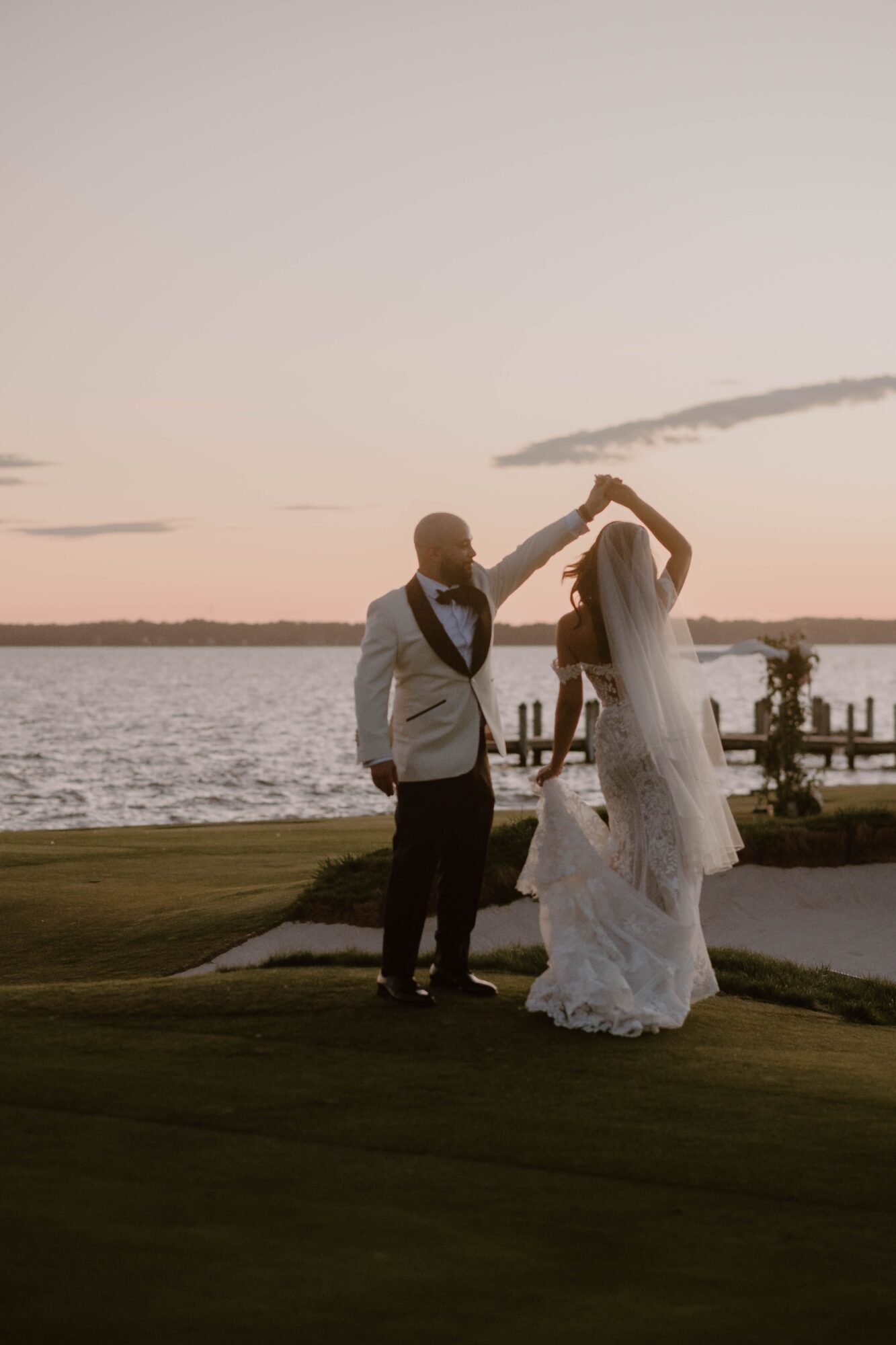 A couple dancing outdoors near a body of water during sunset, with a pier and trees in the background.