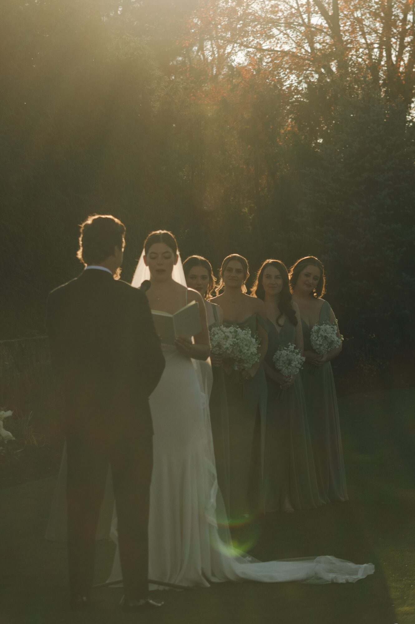 A bride and groom facing a group of four women outdoors, with sunlight filtering through trees in the background.
