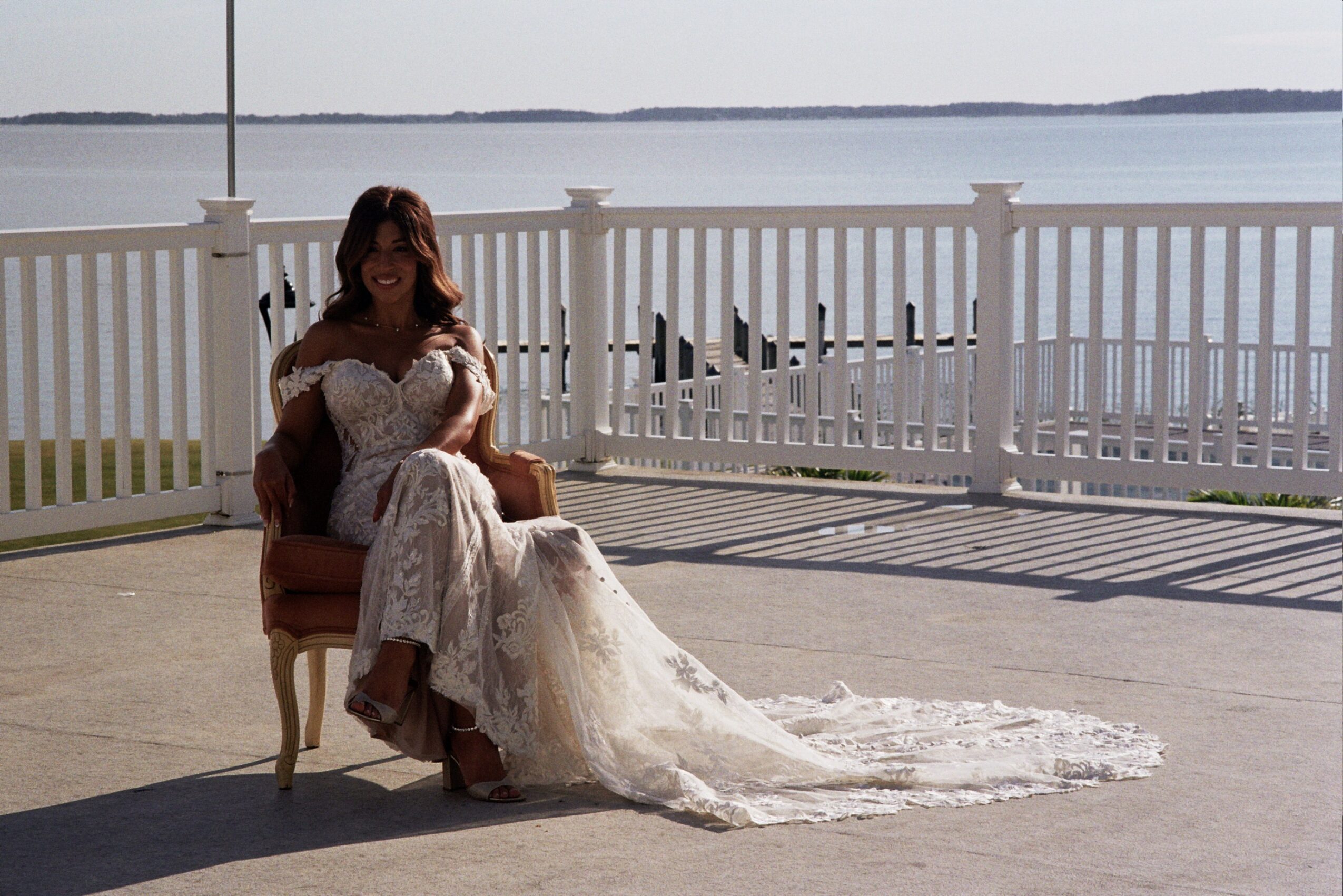 Woman in wedding dress sitting on a chair outdoors near water, with a white fence and boat in background.