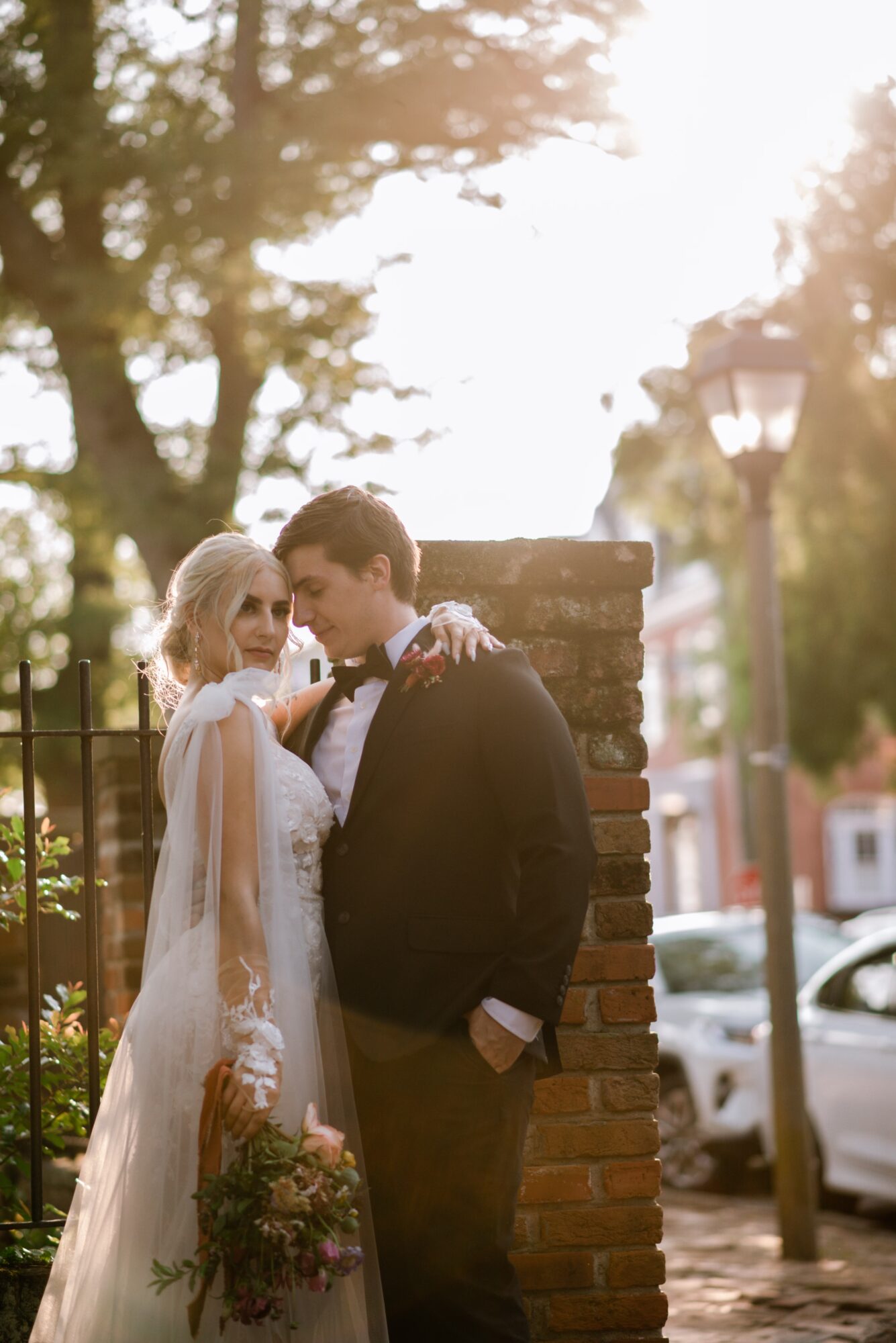A bride and groom stand close together outdoors, with trees and a lamppost in the background.