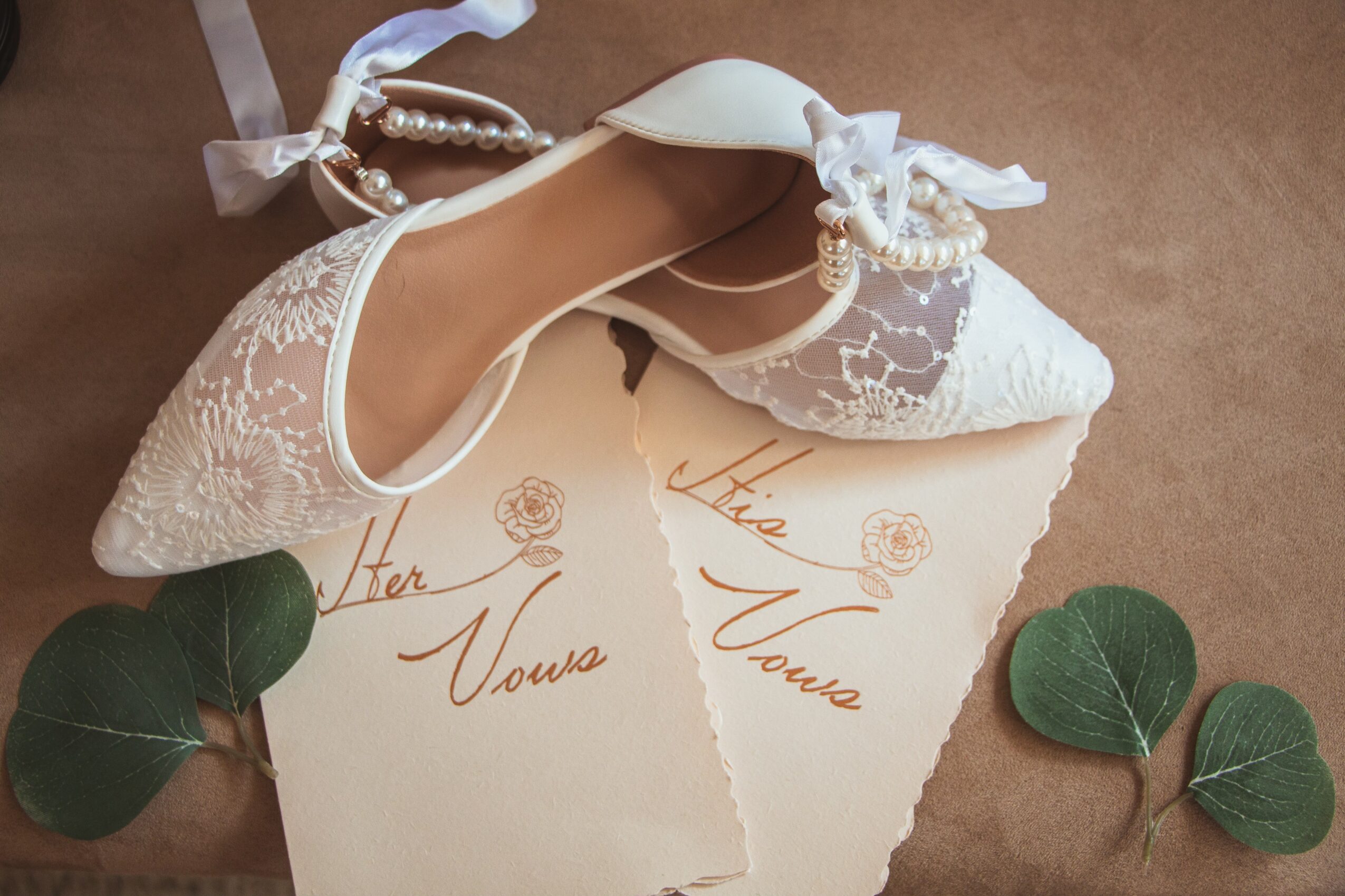 A white lace wedding shoe with ribbon and pearl details, placed on a handwritten note with green leaves nearby.