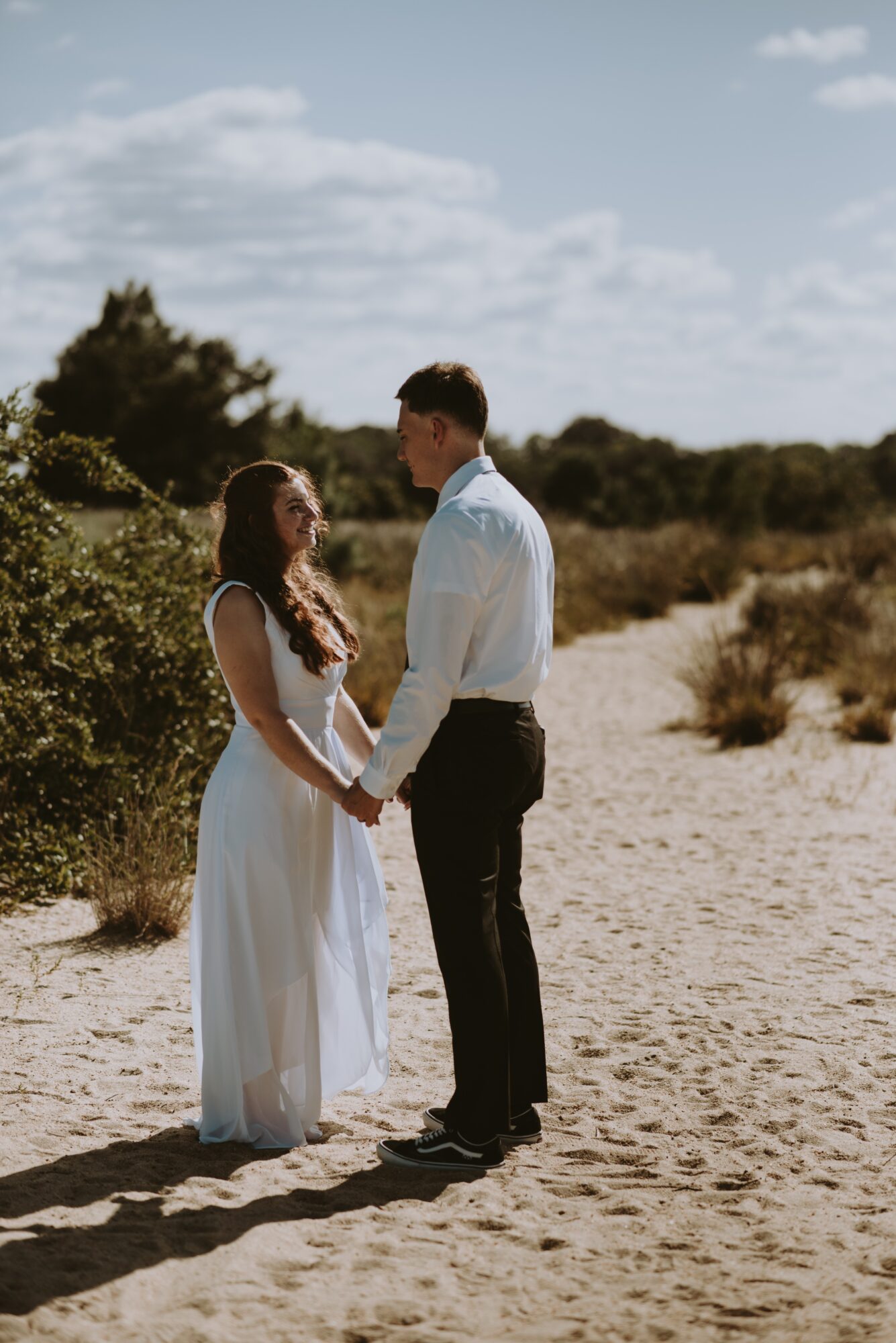 A couple holding hands on a sandy path outdoors, with bushes and trees in the background under a partly cloudy sky.