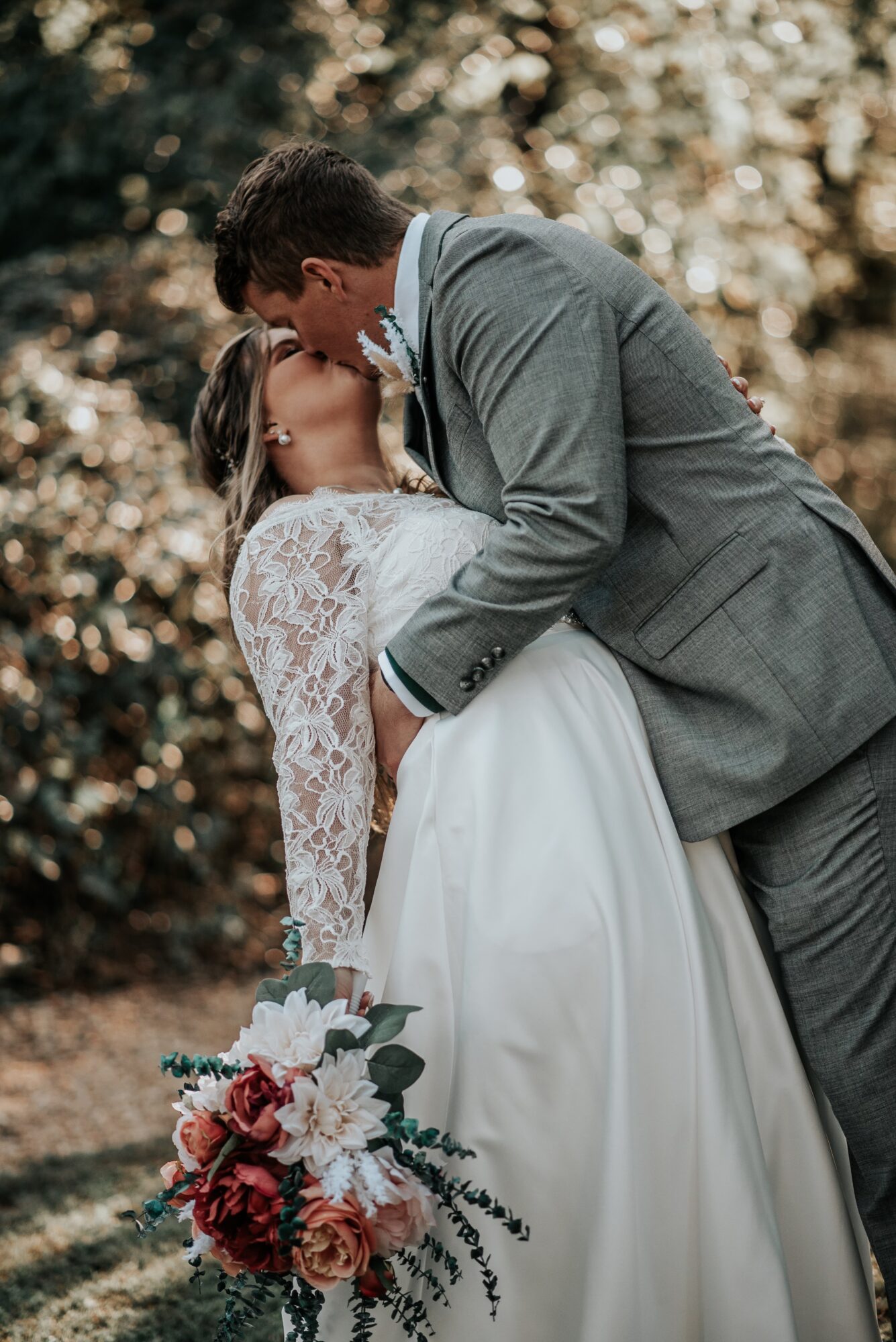A couple in wedding attire sharing a kiss outdoors, with a bouquet of flowers in the foreground.