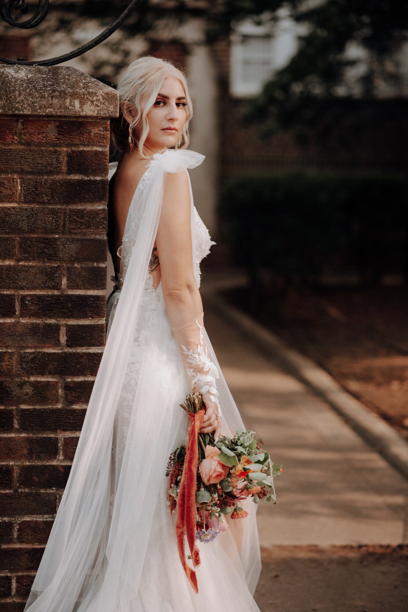 Woman in wedding dress holding a bouquet, standing outdoors near a brick wall, looking to the side.