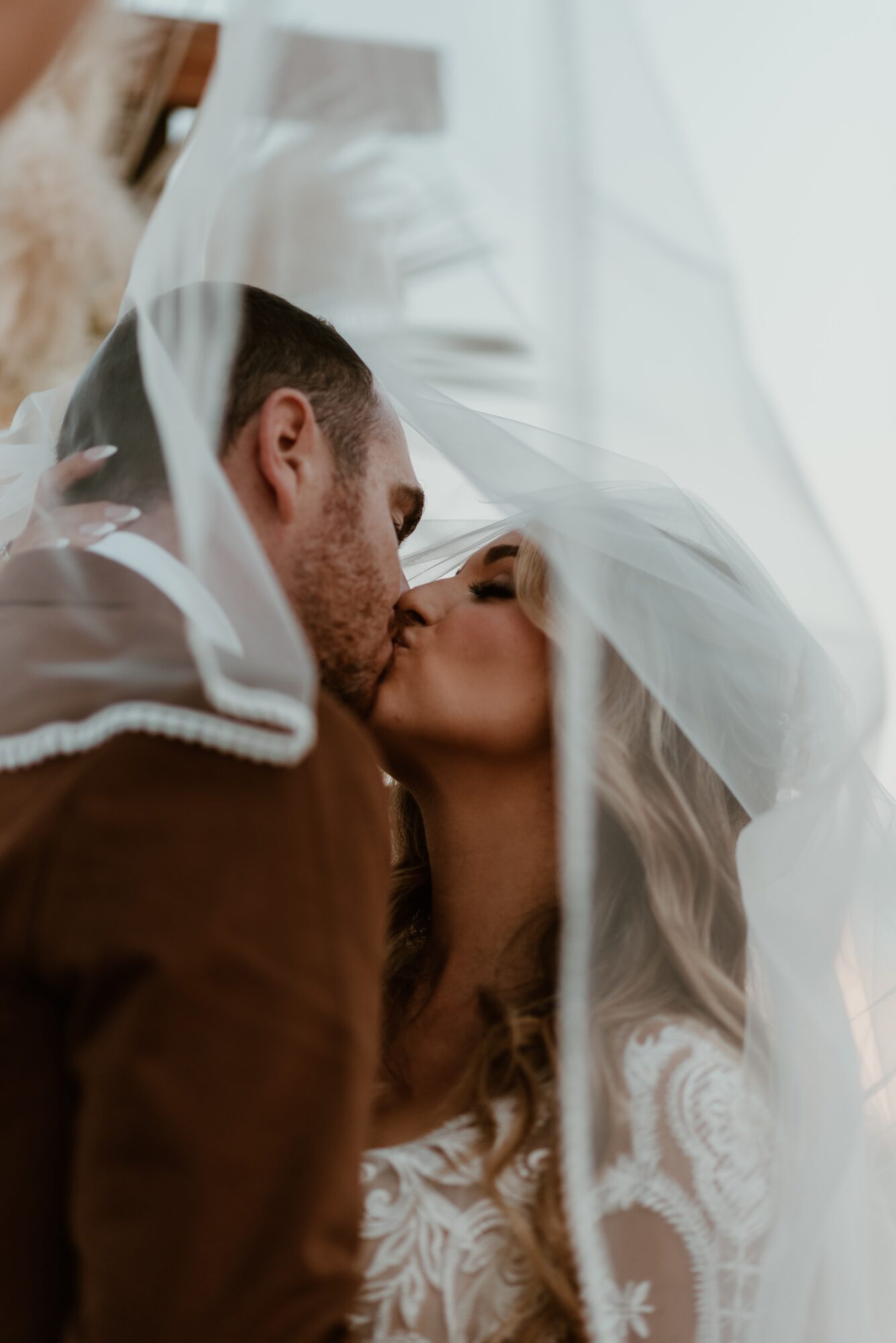 A couple kissing under a sheer veil during a wedding, with soft lighting and lace details on the bride's dress.