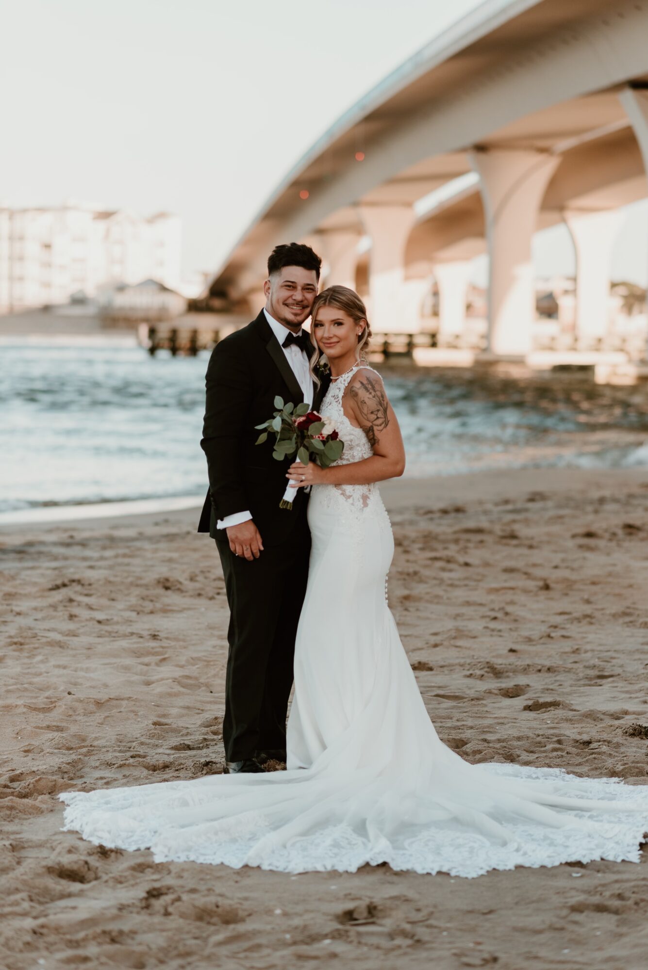 A bride and groom stand on a beach near a bridge, smiling, with the bride holding a bouquet of flowers.