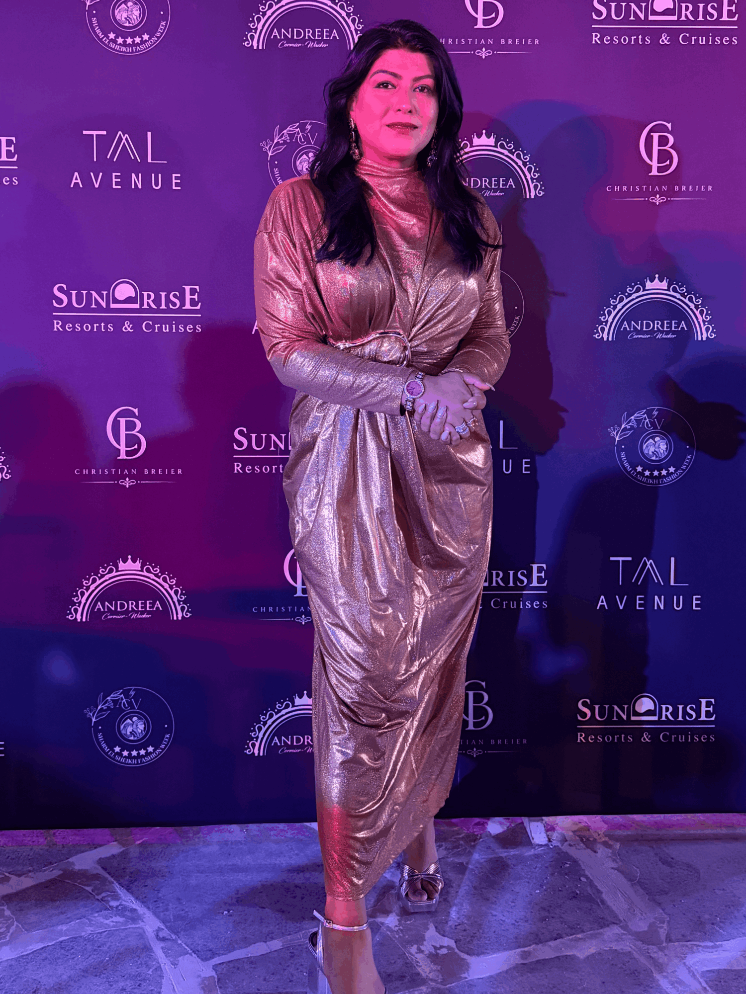 Woman in metallic gold outfit standing in front of a purple backdrop with logos, hands clasped, looking to the side.
