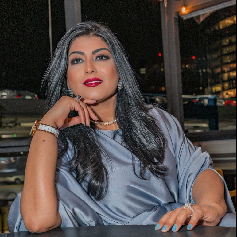 Woman with long dark hair, red lipstick, pearl necklace, sitting indoors at night, looking at camera, wearing a shiny gray top.