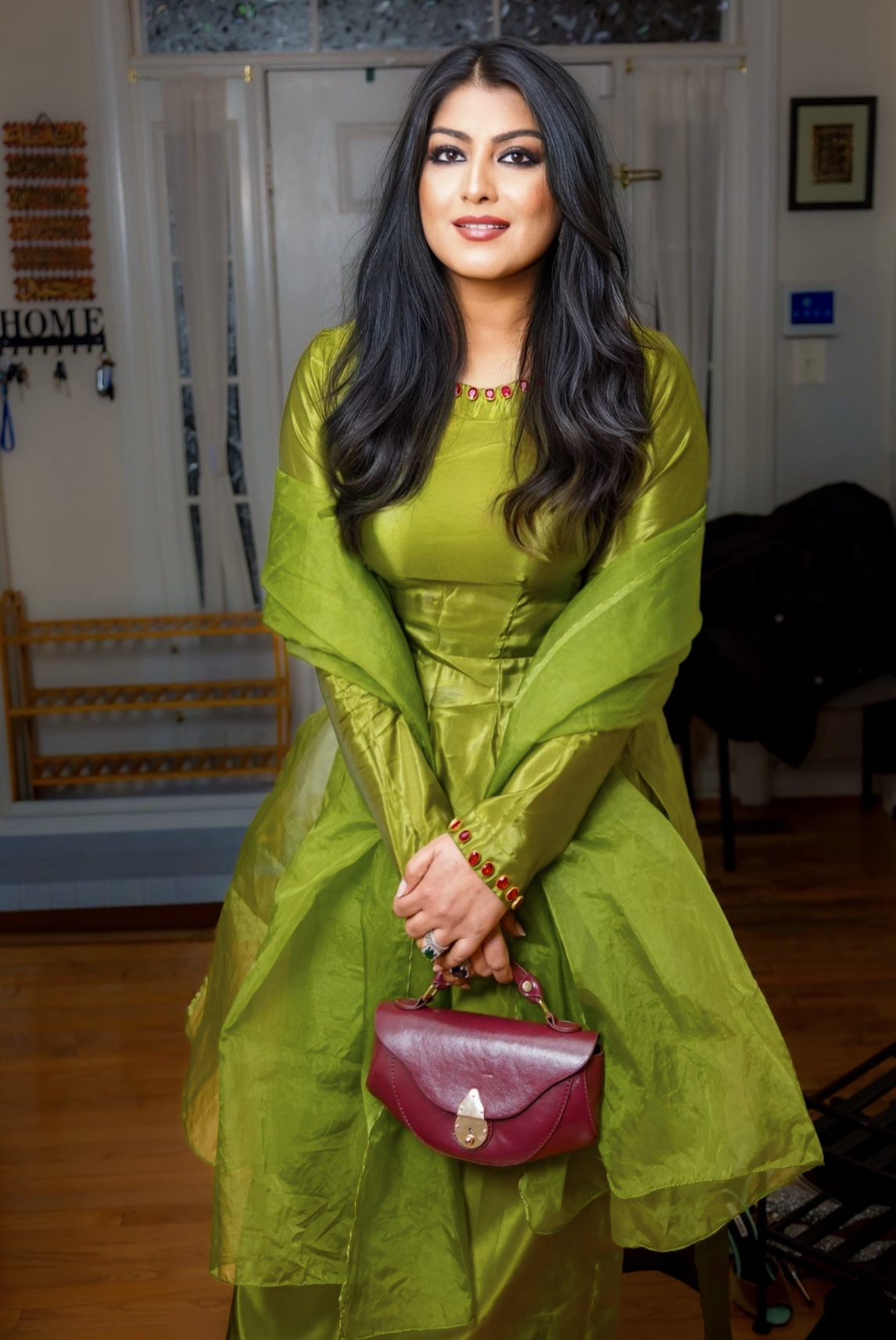 Woman with long dark hair in a green dress holding a red purse, standing indoors in front of a glass door.
