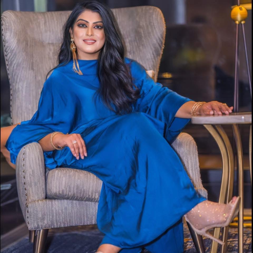 Woman in blue dress sitting on a beige armchair, smiling, with a table and lamp in the background.