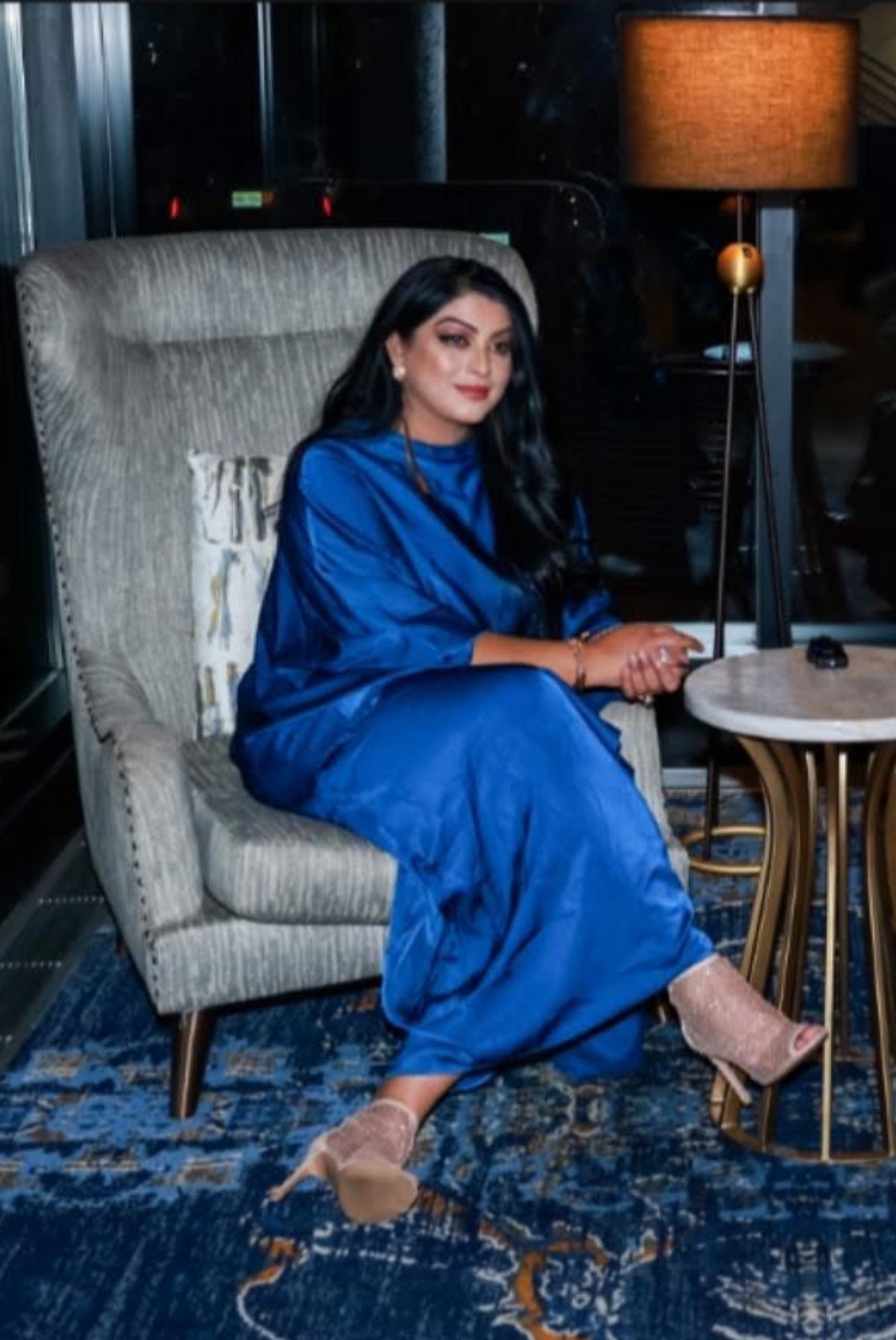 Woman in blue dress sitting on a high-backed chair in a dimly lit room with a lamp and side table.