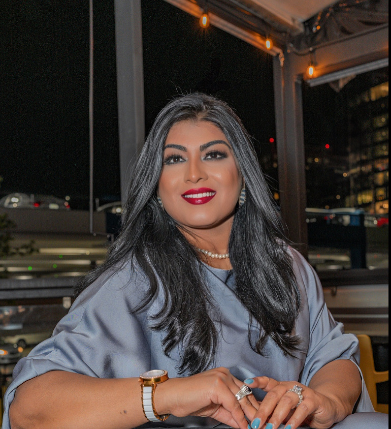 Woman with long dark hair, red lipstick, wearing a gray top, sitting indoors at night with city lights outside.