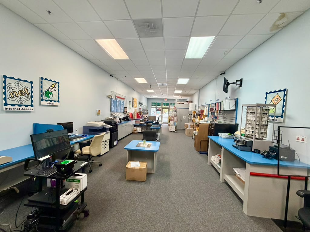 Long room with desks, printers, and office supplies, white walls, ceiling lights, and a window at the far end.