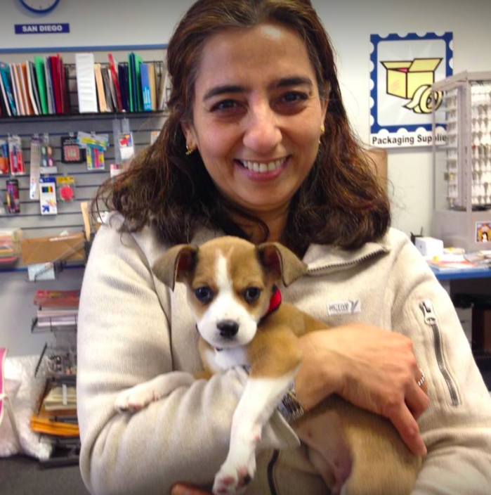 Woman holding a small puppy indoors, smiling, with books and packaging signs in the background.