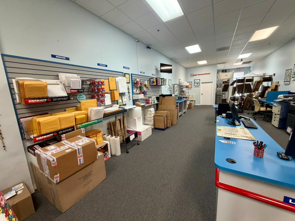 Office supply room with shelves of boxes, paper, and office supplies, and a blue counter with pens and a computer.