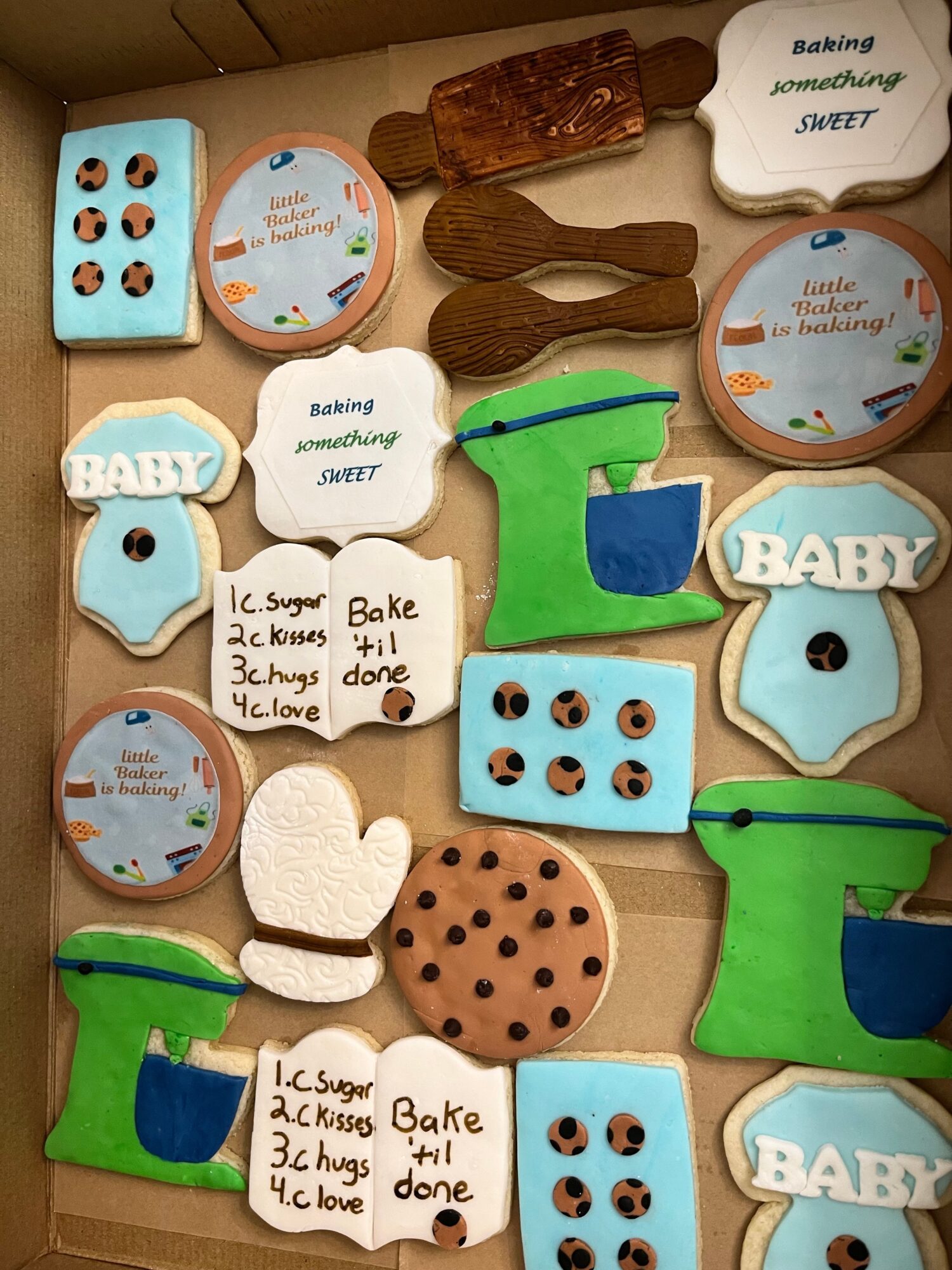 Assorted baking-themed cookies and wooden utensils on a table, including baby-themed cookies, a cookie with chocolate chips, and a note with baking steps.