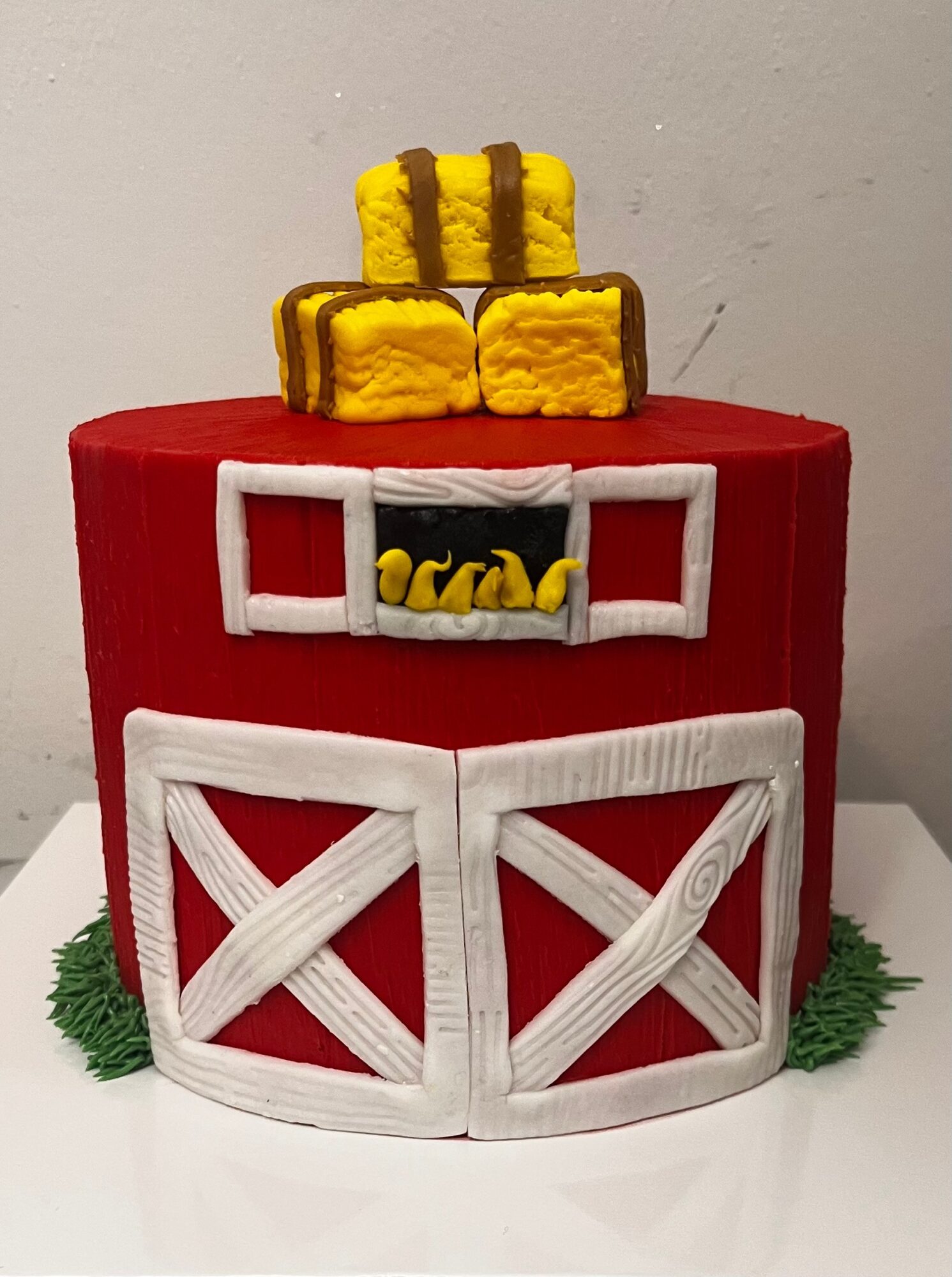 Cake decorated with red, white, yellow, and brown fondant, featuring a barn door, hay bales, and a small window, on a white surface.