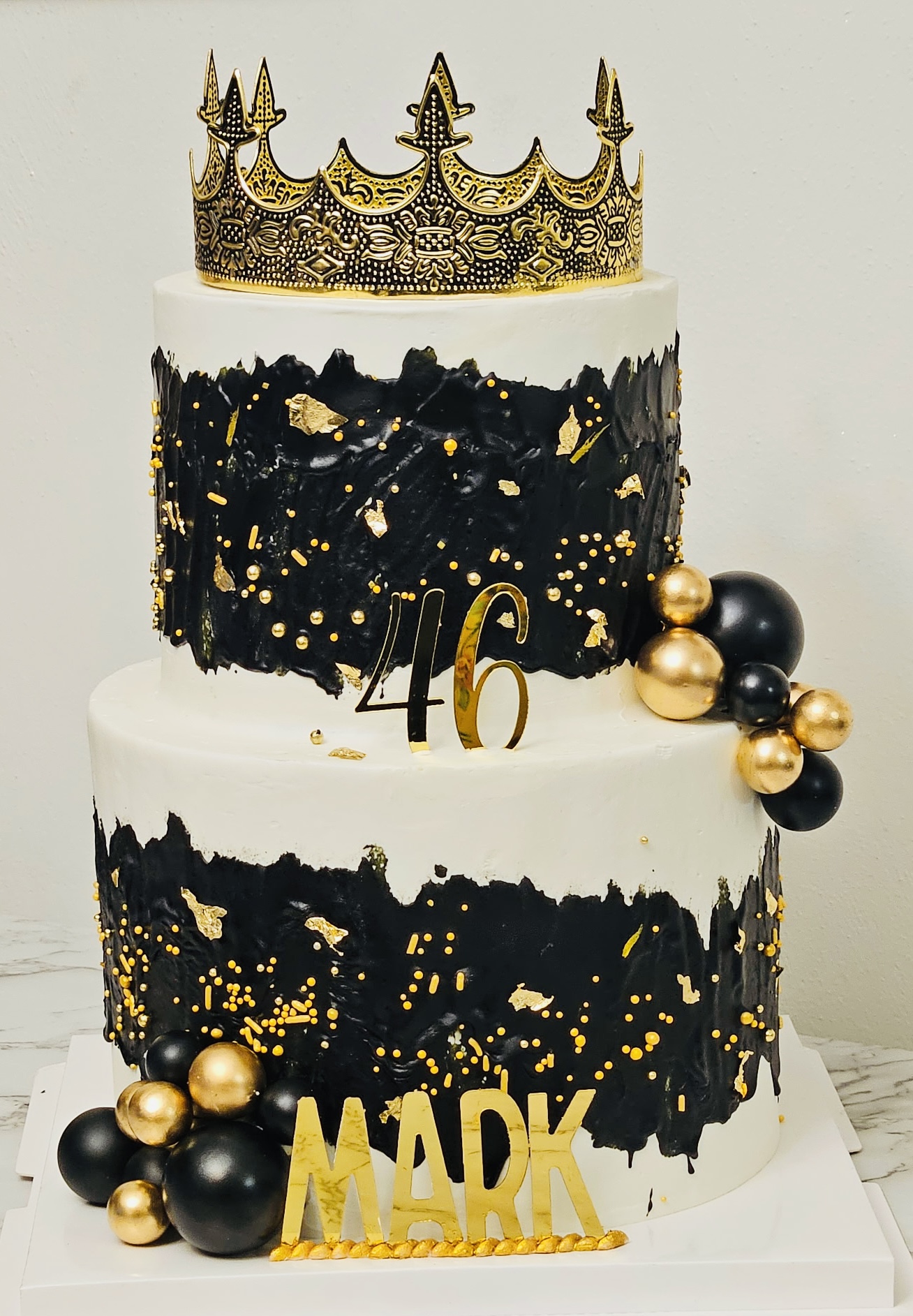 Three-tiered cake with black and gold decorations, gold crown topper, and gold and black accents, celebrating a 46th birthday.