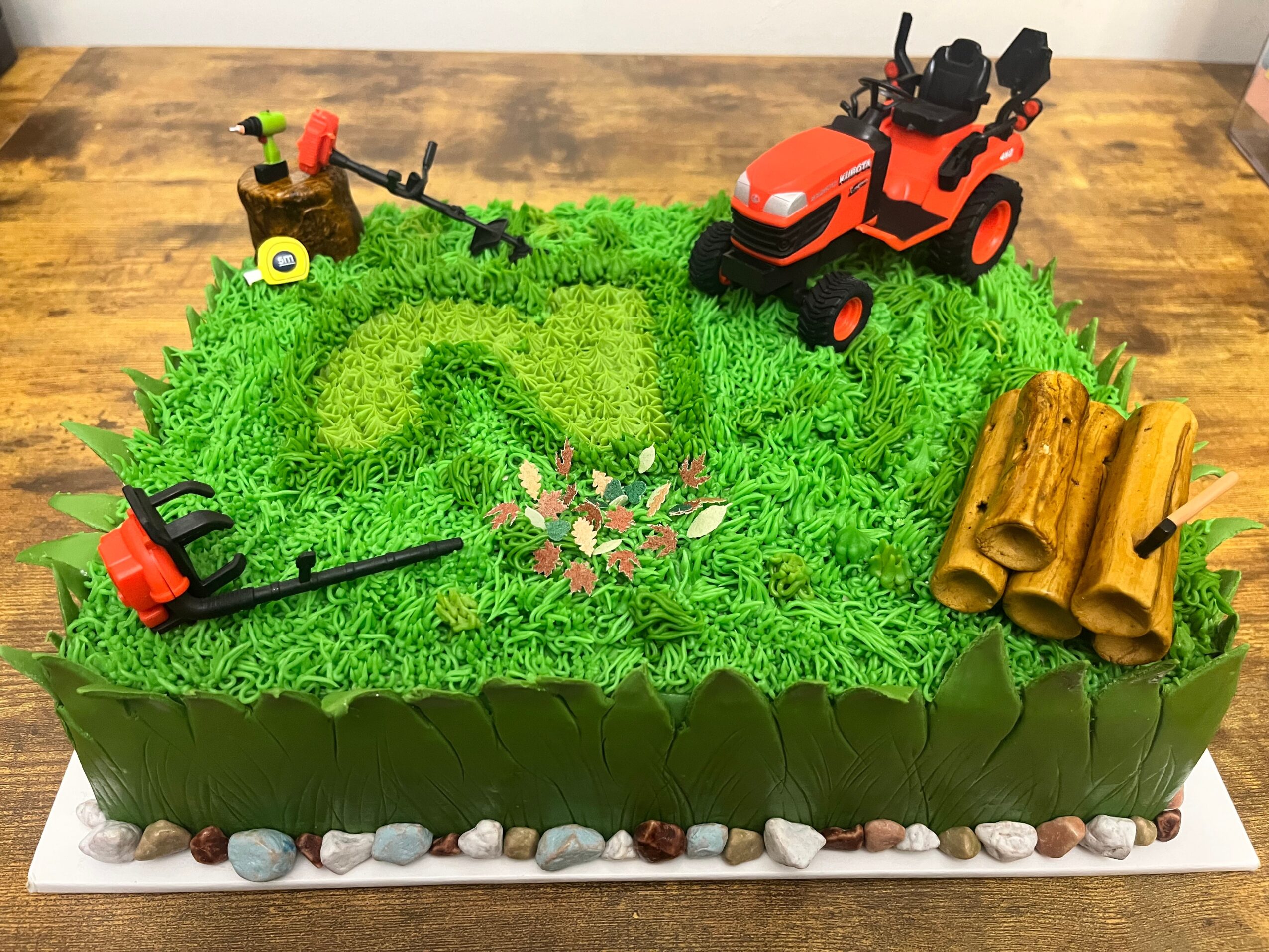 A rectangular cake decorated with green grass-like frosting, small flowers, a red toy tractor, a chainsaw, a pile of logs, and a tree stump with a saw, on a wooden surface.